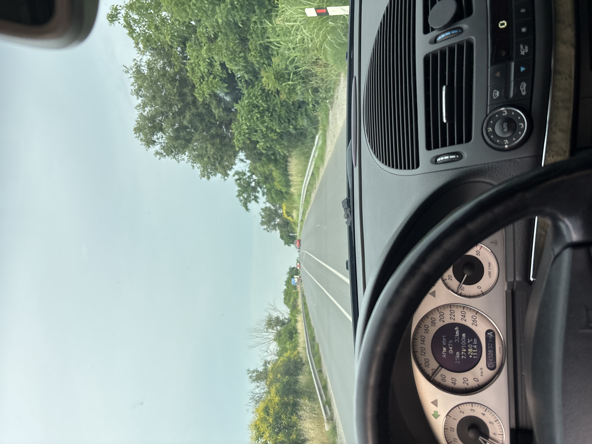 The image is taken from the perspective of the driver of a Mercedes-Benz vehicle, looking forward through the windshield. The car is on a paved road with two lanes in each direction, separated by white lines. The road is bordered by greenery, with trees and bushes on either side. The sky is overcast.
In the foreground, the steering wheel and a portion of the dashboard are visible, including the instrument cluster. The instrument cluster displays the following information:
- Speedometer: indicated speed is between 0 and 40 km/h, with the needle pointing around 20 km/h.
- Tachometer: needle is between 0 and 1,000 rpm, pointing at approximately 800 rpm.
- Digital display:
- "After start": 0:47 h
- 25 km
- 7.7 L/100km
- +28.0 °C
- 111.4 km
- Odometer: 432832 km
In the midground, several cars are visible on the road ahead, traveling in the same direction. There is a guardrail on the left side of the road, and a white and red marker post on the right.
The image was taken in or near Yoakim Gruevo, Bulgaria, as indicated by the metadata. The environment appears to be rural or semi-rural, with vegetation suggesting a natural landscape. The lighting suggests it is daytime, but the overcast sky reduces the intensity of sunlight. There are no people clearly visible in the image. The overall scene depicts a car journey on a road in Bulgaria.