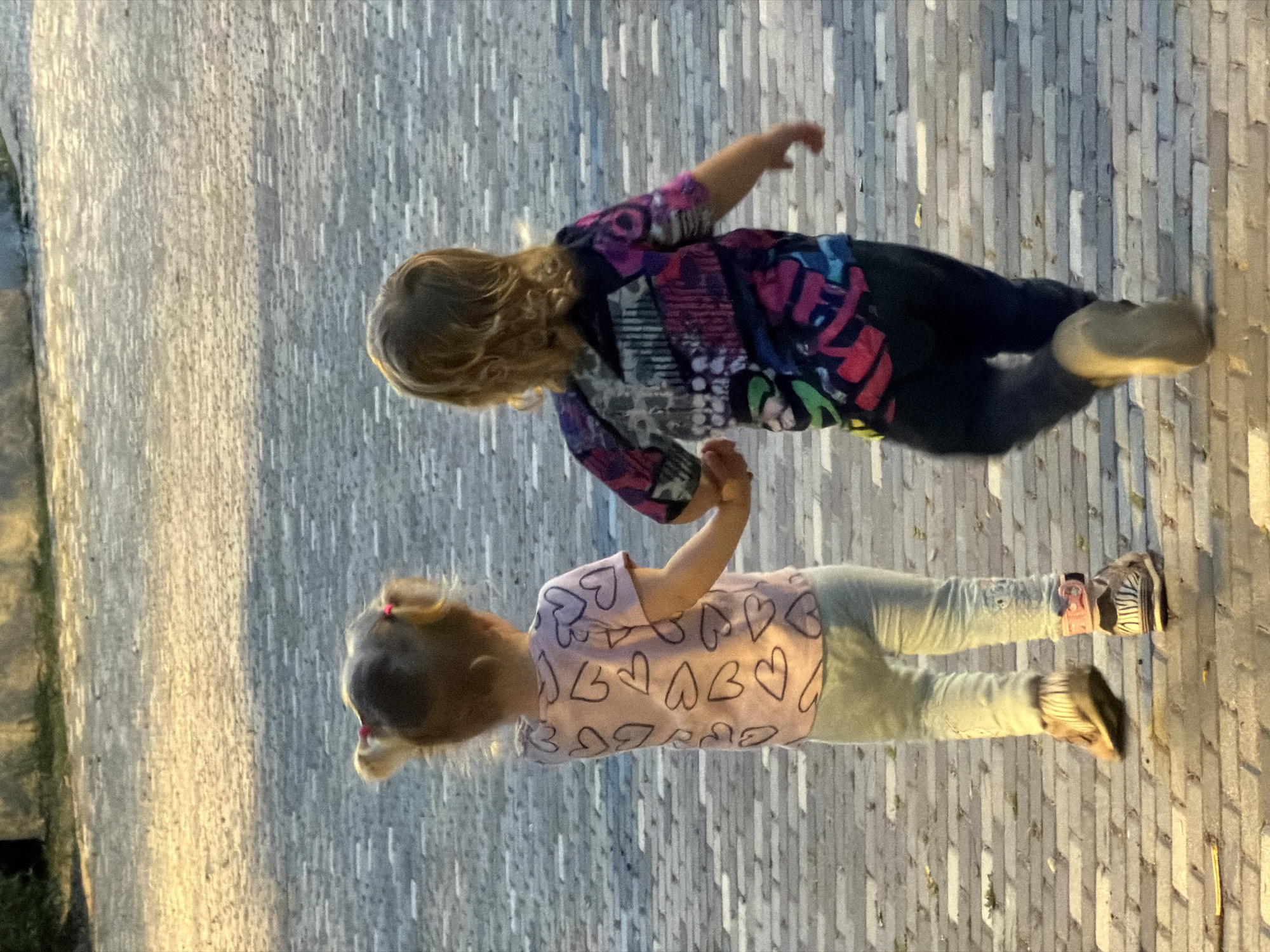 Two young children are walking on a paved walkway, likely in Kiryat Bialik, Israel. The walkway is made of light-colored, rectangular paving stones arranged in rows. One child is wearing a colorful long-sleeved shirt and dark pants, while the other is wearing a light pink shirt with black hearts, light-colored pants, and patterned shoes. The children appear to be moving forward. The lighting suggests it may be late afternoon or early evening, as there are shadows cast on the walkway. There is greenery visible in the background.