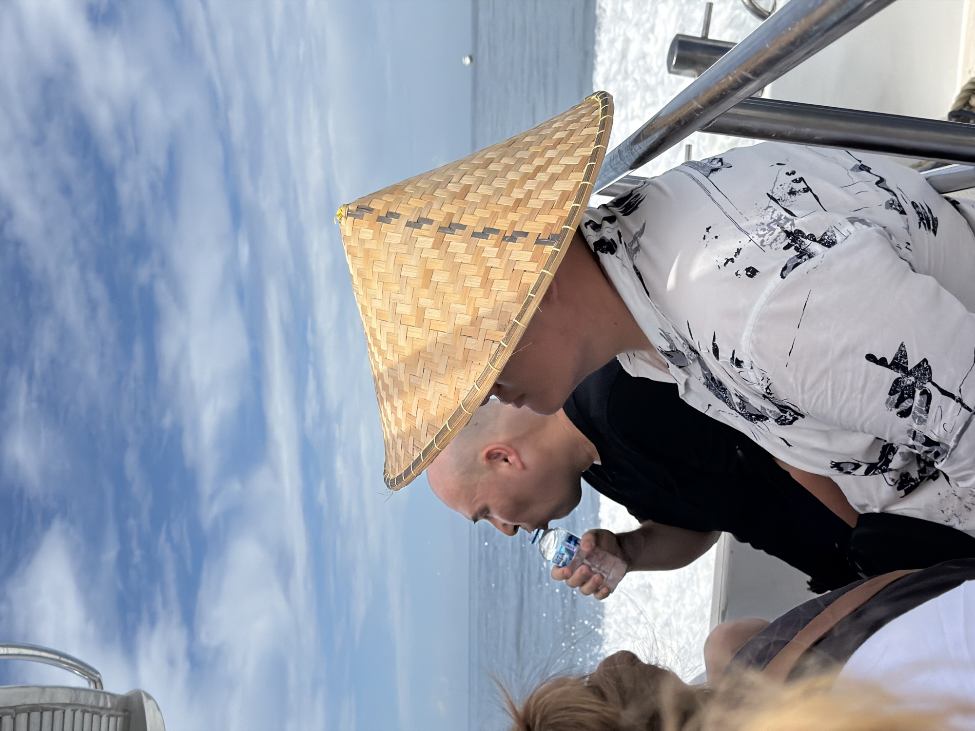 A medium, low-angle shot shows two individuals on a boat during daylight against a background of the ocean and a partially cloudy sky. The person in the foreground, wearing a woven conical hat, is facing right and appears to be drinking from a water bottle. Behind them, another person with a shaved head, wearing a black shirt, is also drinking from a water bottle. The person in the foreground is wearing a white shirt with a dark pattern, and part of another person's hair is visible in the lower part of the frame. Boat railing is visible in the top right. Based on the environment (ocean, cloudy sky) and the presence of traditional conical hats, the scene could be occurring near Denpasar, Indonesia.
