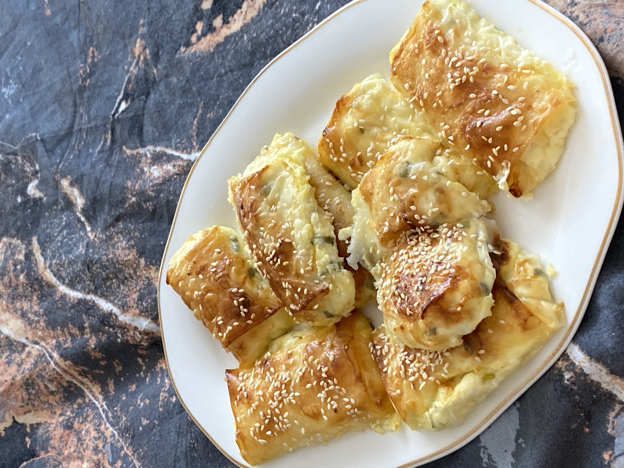 A top-down shot shows a white platter piled with baked pastries, likely bourekas, adorned with sesame seeds on top, against a textured background. The pastries appear golden-brown and puffed, suggesting a flaky crust. The platter has a thin gold rim. The background has a dark, marbled appearance, potentially a countertop or table surface. No people or actions are visible in the image. The location of Chaikovsky, Russia is not apparent from the visual information provided.