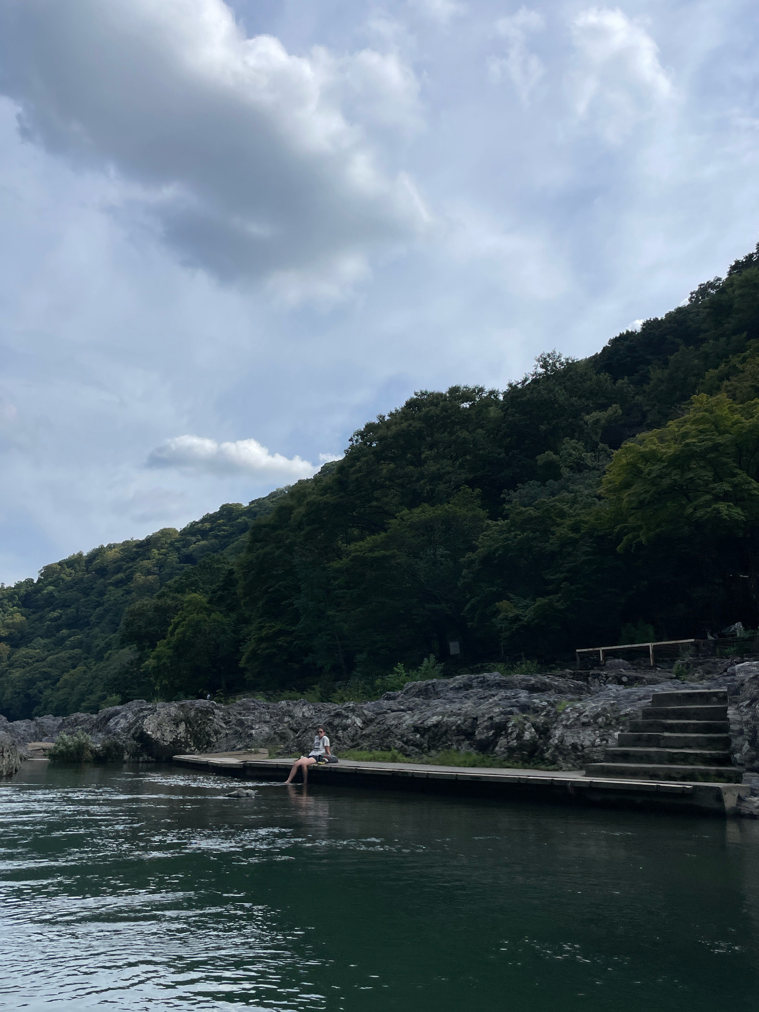 A person is sitting on a wooden platform next to a river in Arashiyama, Kyoto. The person is wearing a white t-shirt, blue shorts, and sunglasses. They have their feet in the water. The river is dark green and calm, with ripples on the surface. Behind the platform, there are large grey rocks and a steep, tree-covered hillside. The trees are dense and dark green, with some lighter green foliage mixed in. The sky is cloudy with patches of blue showing through. To the right of the platform, there is a set of concrete stairs leading up the rocky slope. There is a wooden railing at the top of the stairs. The overall scene is serene and natural.