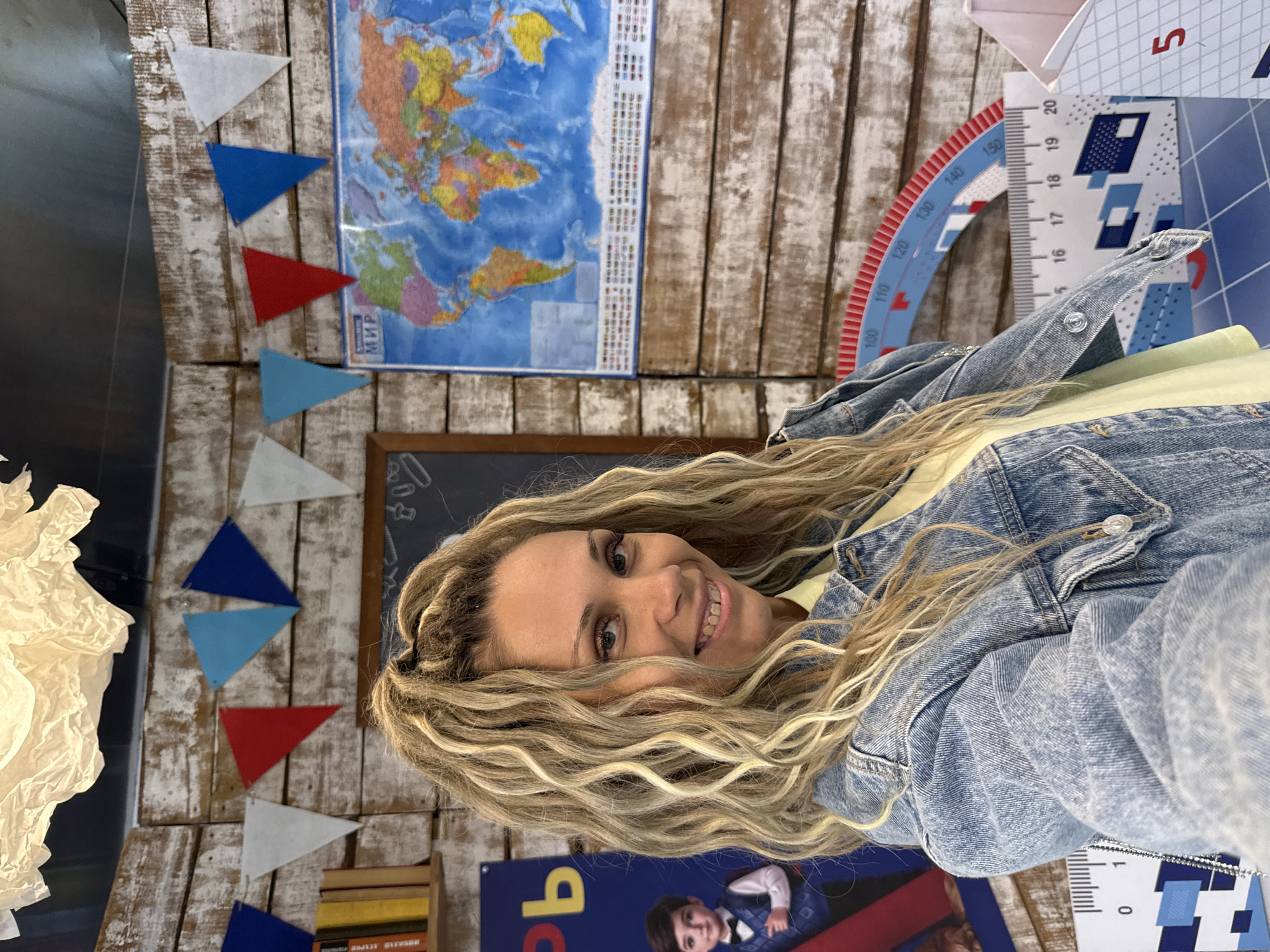 A woman with blonde, curly hair and a denim jacket smiles directly at the camera. She is in a room decorated with a world map, a chalkboard, a shelf with books, and colorful triangular flags strung across a wooden wall. In the foreground, there are measuring tools and white boxes with red numbers, suggesting an educational or child-friendly setting. The background also features a poster of a young boy in formal attire. The scene appears to be indoors, possibly a classroom or a themed play area. The specific location in Ignatovo, Russia cannot be determined from the visual information provided.