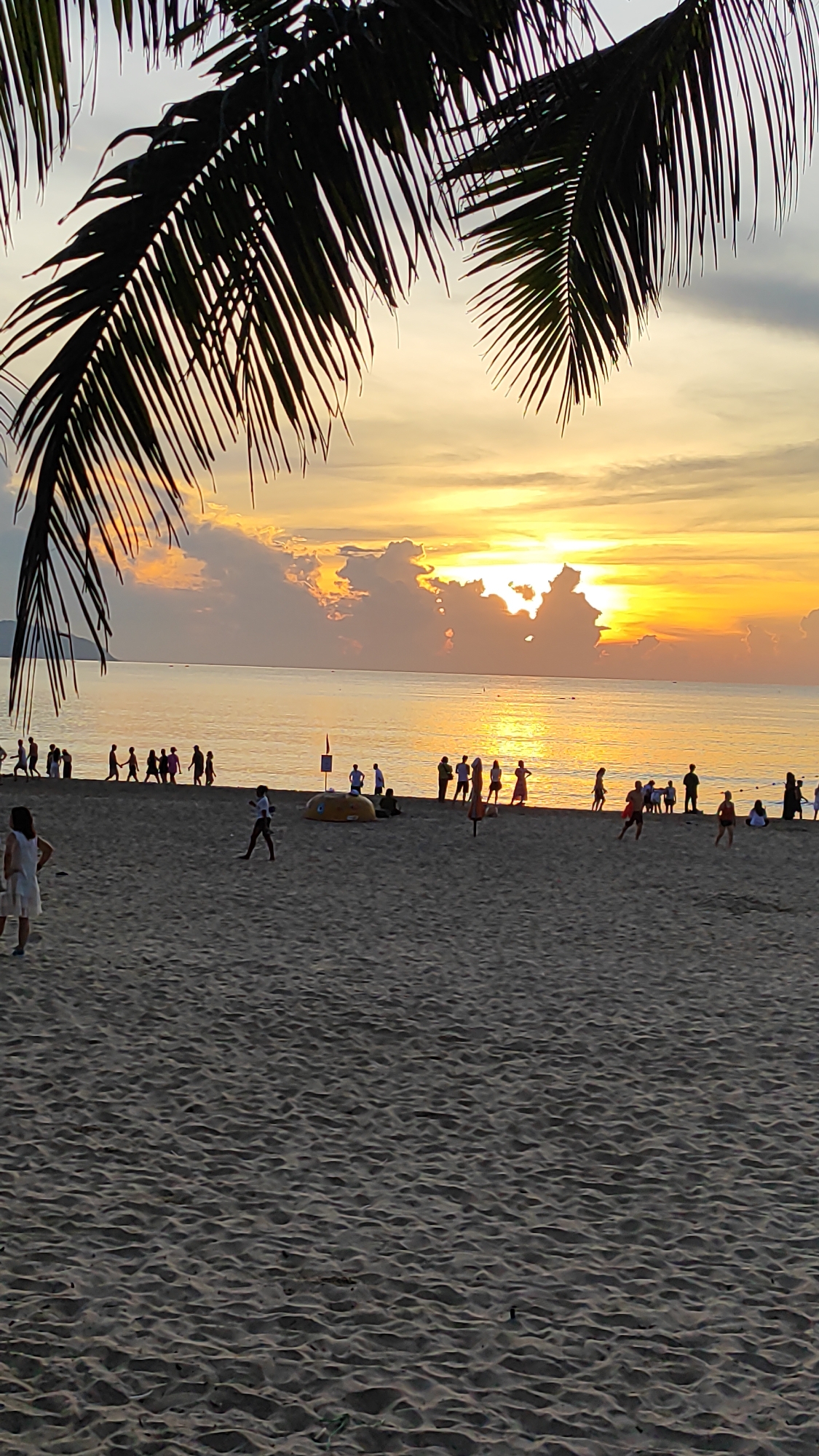 This is a full shot of a beach scene at sunset in Da Nang, Vietnam, viewed from the sand with palm fronds in the foreground. The sun is setting over the ocean, casting a golden glow across the sky and water. Numerous clouds are illuminated by the setting sun, creating a dramatic backdrop. A line of people are walking along the shoreline, some appearing to be heading towards or away from the water. Several individuals are spread across the sandy beach, with some playing and others observing. In the middle ground, there are a few objects including what appears to be a yellow inflatable raft and a red flag on a pole. The sand is textured with small ripples, indicating it is a typical beach environment. The overall atmosphere is one of a leisurely evening by the sea.