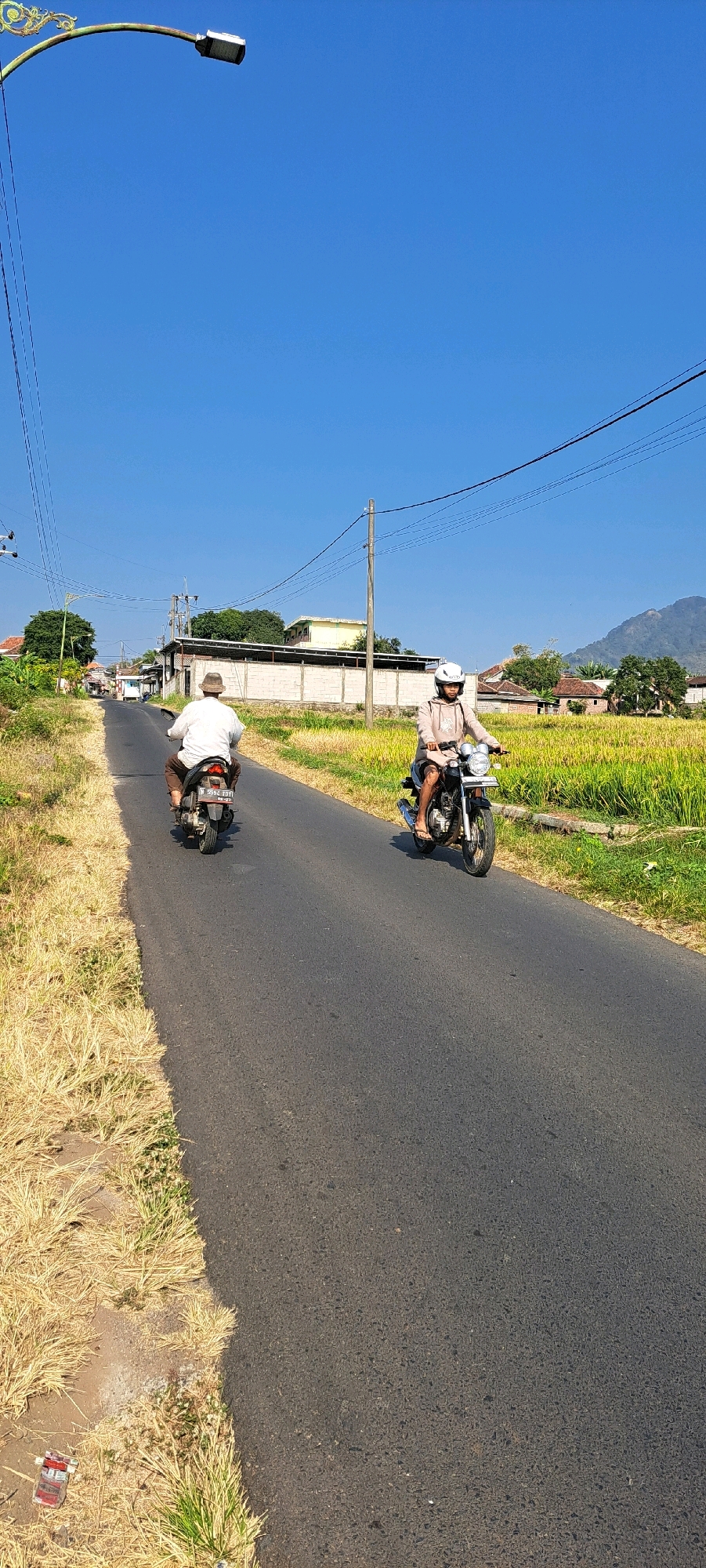 On a clear, sunny day in Sumber Gedang, Indonesia, two individuals are riding motorcycles on an asphalt road. The road is flanked by dry, light-brown grass on the left and a vibrant green rice paddy on the right. In the background, there are houses and buildings, as well as power lines stretching across the blue sky. A mountain range is visible in the far distance.
The person on the left, riding a black motorcycle, is seen from the rear, wearing a white shirt and brown pants, and a hat. The motorcycle has a license plate that reads "2594 FBY". The person on the right, riding a darker motorcycle, is wearing a light-colored hooded sweatshirt and shorts, and a white helmet. This individual is facing forward, and the motorcycle has its headlights on.
The scene appears to be a rural or semi-rural area. The overall impression is one of a typical day in the Indonesian countryside, with the focus on the two motorcyclists and the surrounding landscape.
