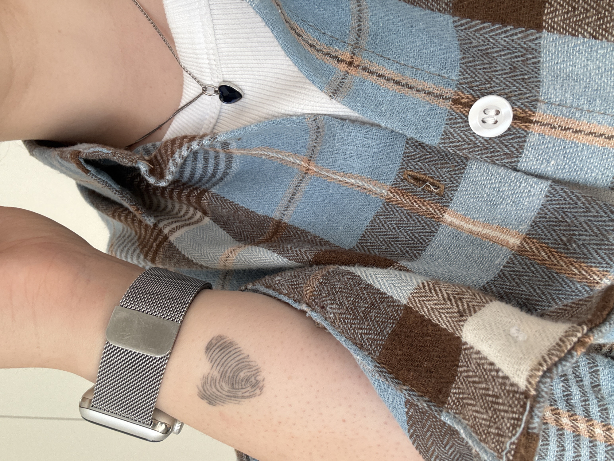 The image shows a close-up view of a person's left arm and shoulder, wearing a blue and brown plaid shirt over a white ribbed tank top. A silver mesh watch band is visible on the wrist. On the upper arm, there is a tattoo of a fingerprint in the shape of a heart. A silver necklace with a dark blue heart-shaped pendant rests on the person's collarbone. The background is a plain white wall, suggesting an indoor setting. There is no indication of the location being in Lyubertsy, Russia.