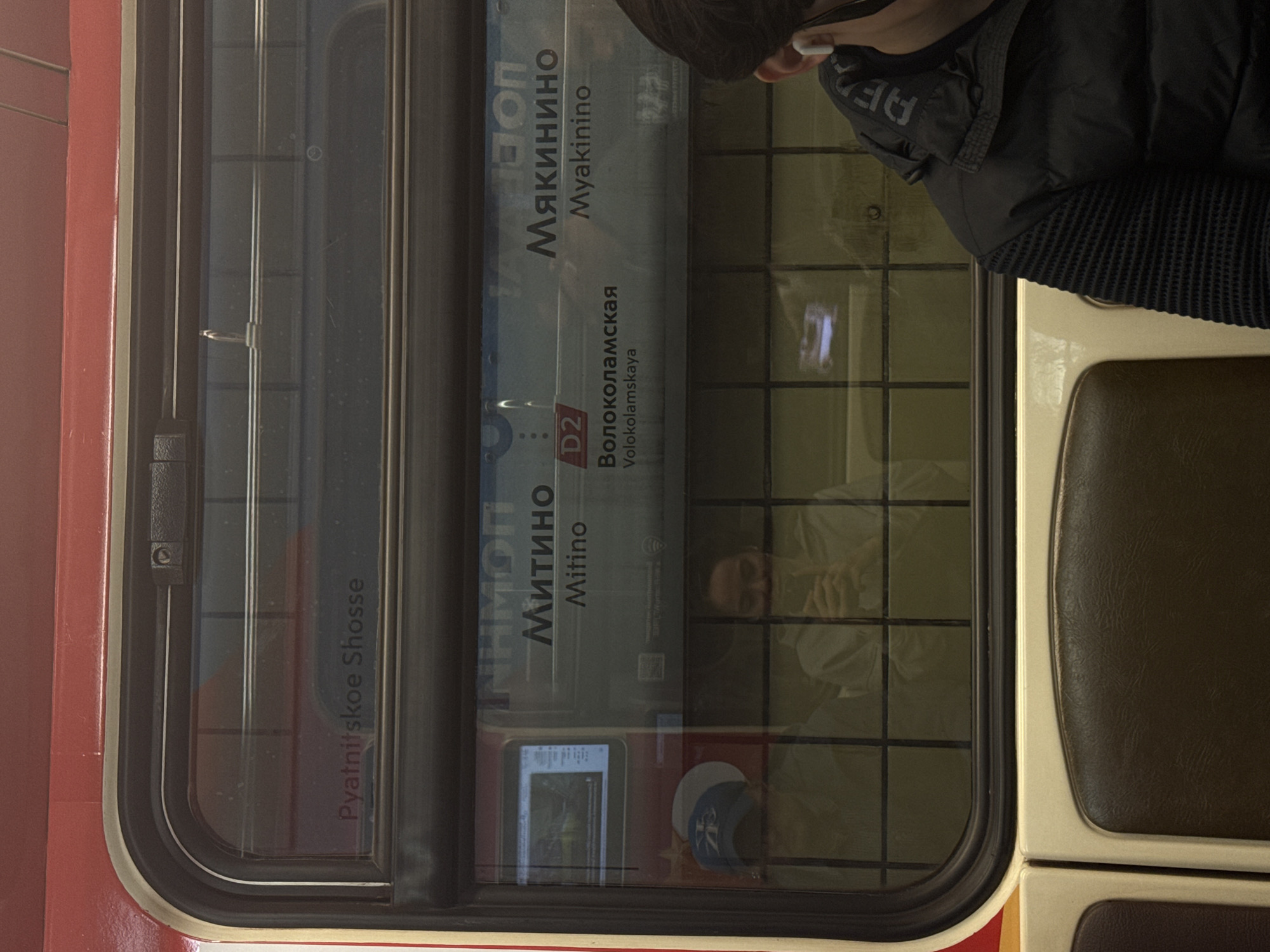 The image shows the interior of a Moscow Metro train car. The window displays station names in Cyrillic and Latin script, including "Mitino" (Митино), "Volokolamskaya" (Волоколамская), and "Myakinino" (Мякинино). The train line "D2" is also visible. A person wearing a dark jacket is seated next to the window. Reflections in the window show passengers and objects in the train car. The interior includes a seat with brown and beige upholstery.