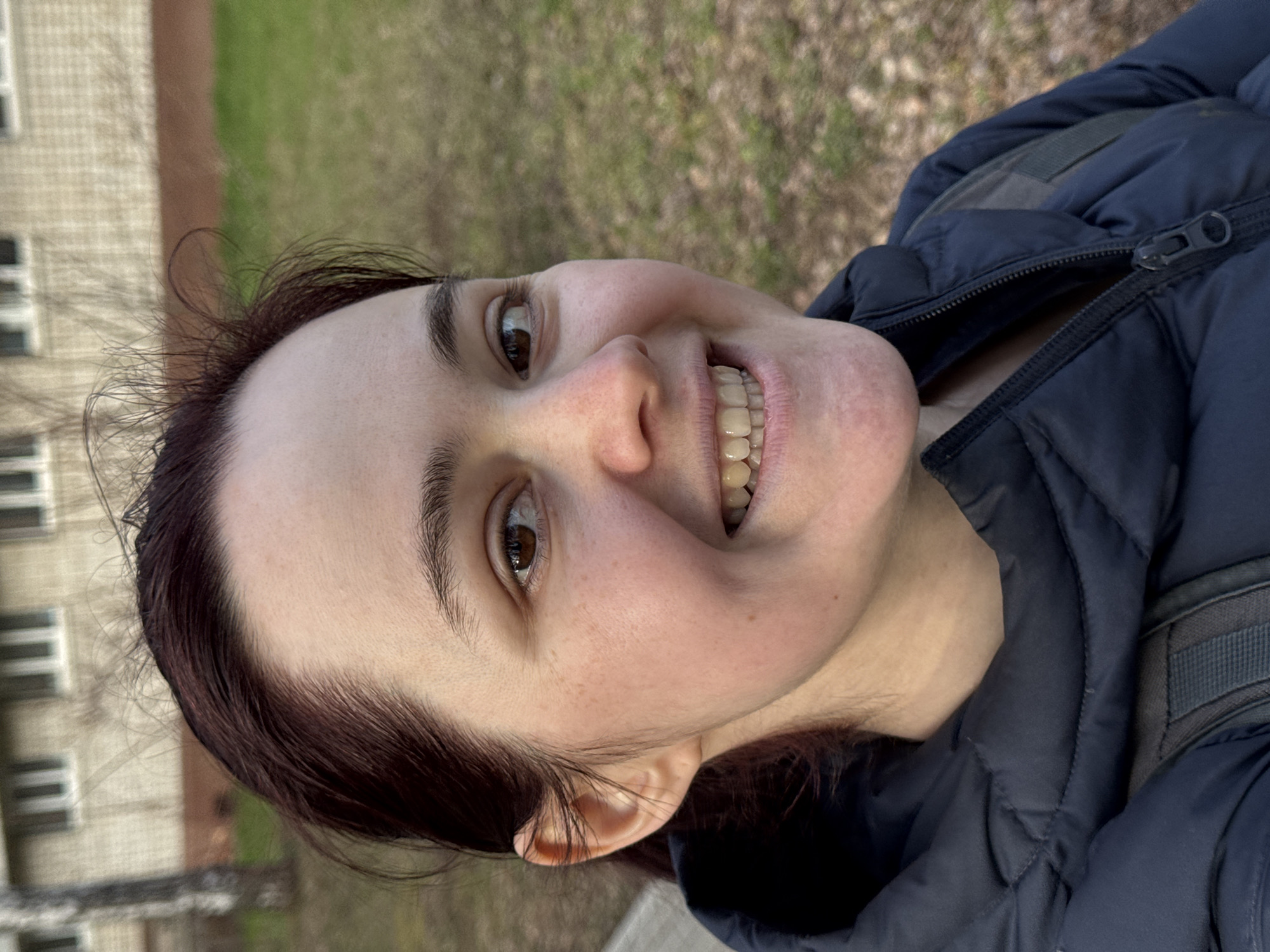 The media file displays a close-up, outdoor portrait of a young to middle-aged woman from Izhevsk, Russia, captured in daylight. She has fair skin with visible freckles, dark brown hair, and brown eyes, and is looking towards the viewer with a broad, cheerful smile that shows her teeth. Her expression conveys happiness and friendliness.
The woman is wearing a dark blue, padded or puffer jacket, with a visible zipper detail on the chest. A strap of what appears to be a backpack is visible over her right shoulder, suggesting she is out for a walk or engaged in an outdoor activity.
The background, visible behind and around her, is softly blurred but shows an outdoor setting. To her left, part of a light-colored building with multiple rectangular windows can be seen, likely an apartment block or institutional building. To her right and further behind, there is green ground cover, possibly grass, interspersed with some bare branches or dry foliage, hinting at either early spring, late autumn, or a mild winter day.
The lighting is natural and diffuse, without harsh shadows, suggesting an overcast day or an evenly lit outdoor environment. There is no visible text in the image.