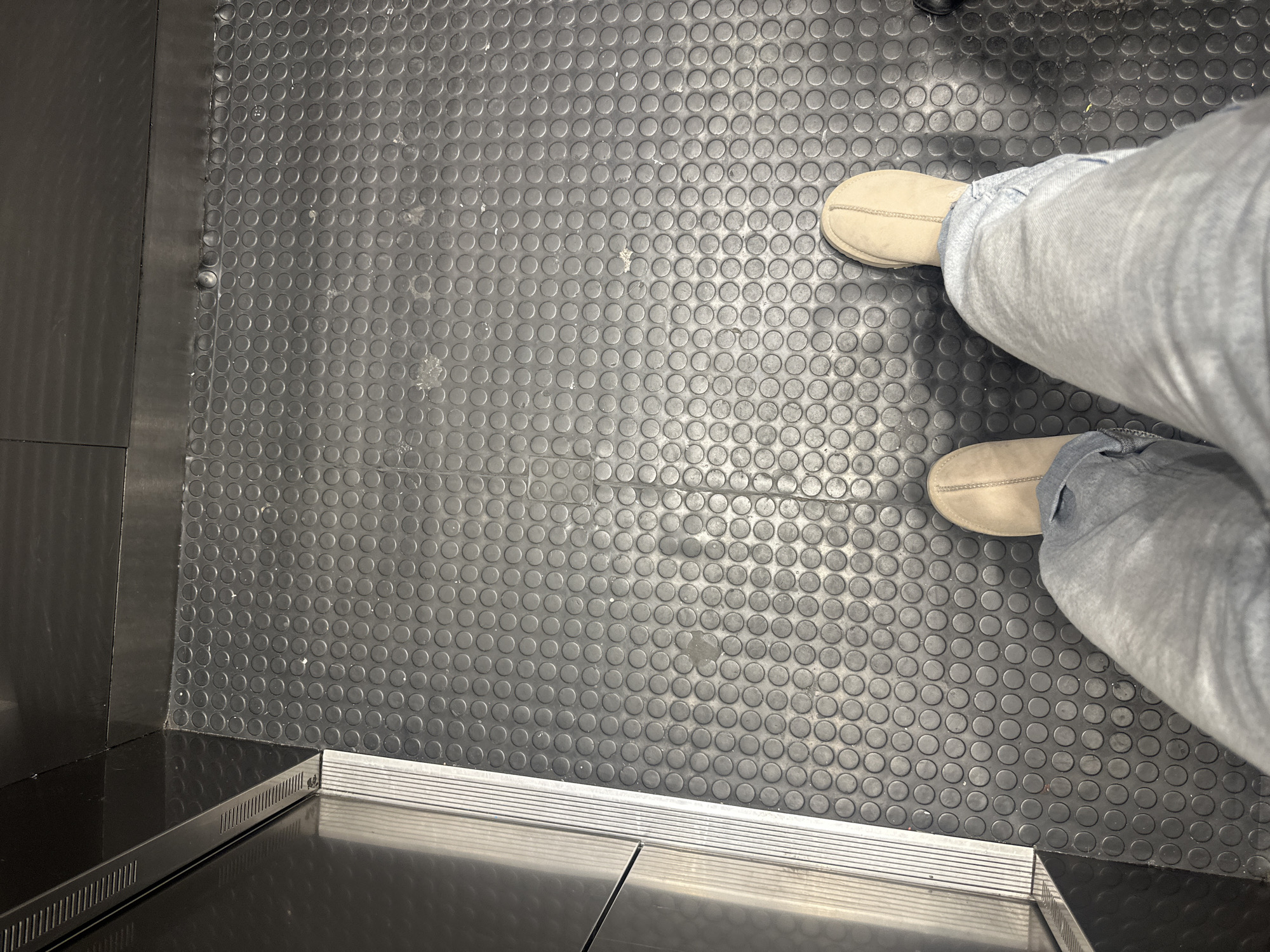 The image shows a person standing in what appears to be an elevator. The floor of the elevator is covered in black rubber with a repeating circular pattern. The person is wearing light-colored slippers and blue jeans. The elevator has a metal door and trim, and the overall lighting is somewhat dim. It is not possible to identify the location as Mexico City based on the visual information provided.
