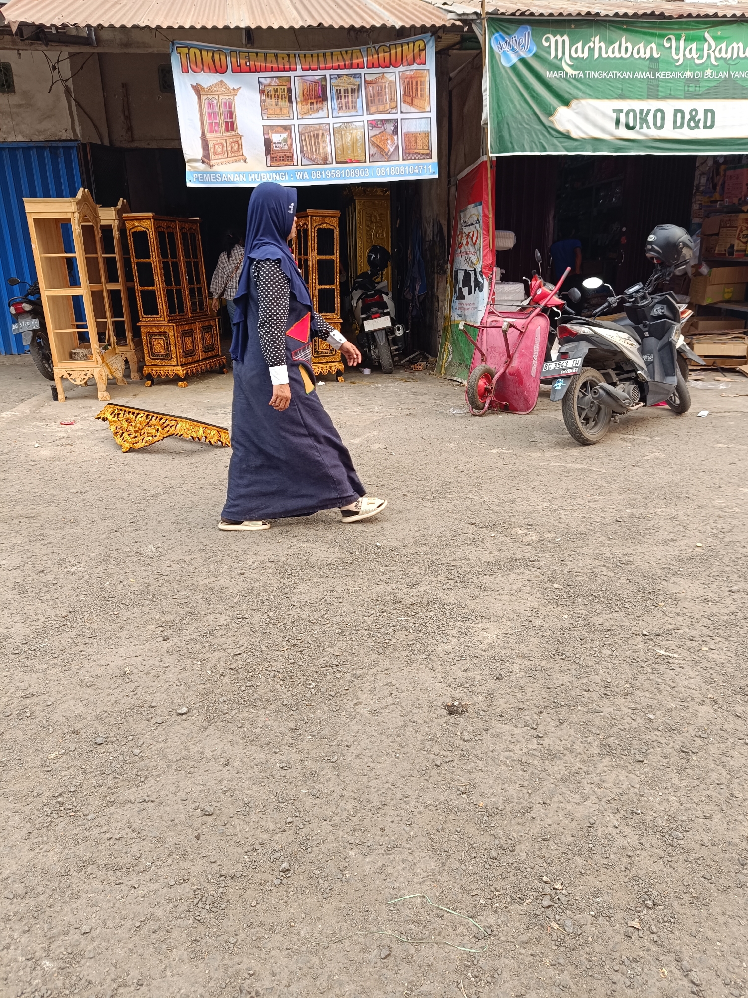 An eye-level, medium shot captures an outdoor scene in front of a shop selling furniture in Kayu Agung, Indonesia. A woman wearing a dark blue hijab and a long, dark blue dress with a black and white polka dot top walks from left to right, her body angled away from the camera. She is wearing light-colored shoes. To her left, several ornate wooden furniture pieces are displayed, including tall, empty wooden shelves and detailed golden cabinets. A golden carved wooden piece lies on the ground in front of her. Behind the furniture, a banner with text is visible, likely advertising the shop. To the right of the woman, a red wheelbarrow and a black scooter are parked near the entrance of another shop, identified by a green banner with Indonesian text. The ground is covered in gravel and dirt. In the background, more scooters and shop fronts are visible.