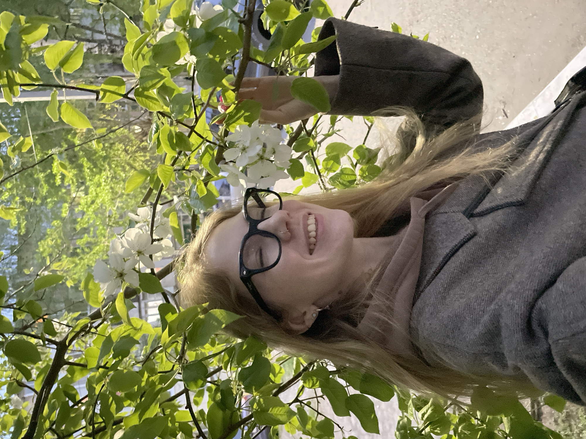 A young, smiling woman with long blonde hair, wearing black-framed glasses, a light pink top, and a gray coat, stands amongst the blossoming branches of a tree. The tree has green leaves and white flowers. The background suggests an urban environment, possibly a park or garden in Izhevsk, Russia, given the user's prompt. The photograph appears to have been taken from a slightly low angle. The woman's right hand is visible near the flowers.