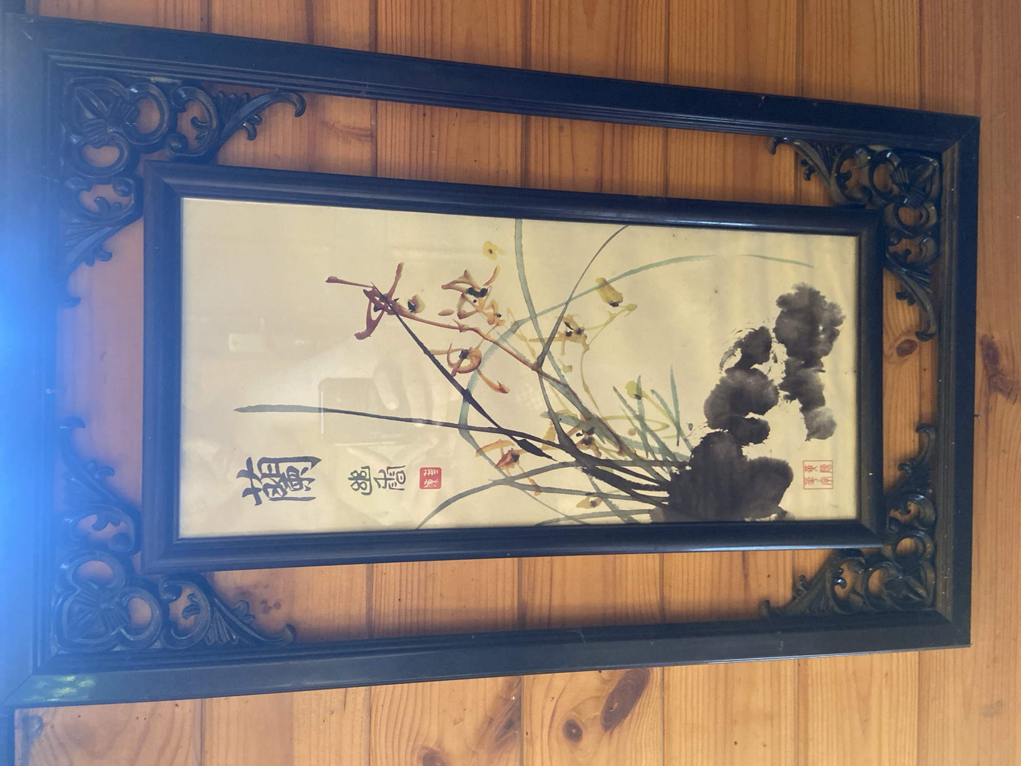 A framed watercolor painting of orchids hangs on a wooden paneled wall. The painting depicts several delicate orchids with yellow and brown petals against a pale background. Dark ink washes are used to create foliage and a decorative seal. The frame is dark wood with ornate, black decorative corner pieces. The image is a head-on, eye-level shot of the artwork, with no people visible. The overall scene is static and indoors. There is no information in the image to suggest its location in Kazan, Russia.