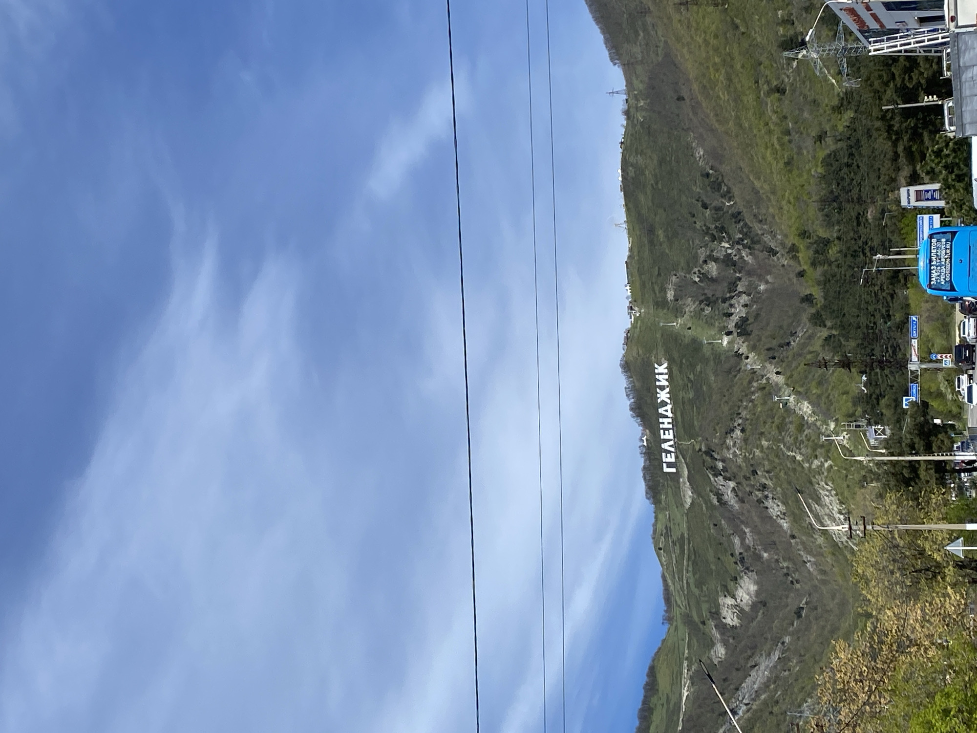 A panoramic landscape photograph captures a large, green mountain under a wide expanse of clear blue sky with a few wispy white clouds. The image is taken during the daytime, suggesting fair weather.
Prominently displayed on the upper slope of the mountain are large, white Cyrillic letters spelling "ГЕЛЕНАДЖИК" (Gelendzhik), indicating the location in Russia. The mountain itself is covered in sparse green vegetation, with some areas appearing rockier or less densely forested.
Stretching horizontally across the middle of the frame are several dark, parallel lines, likely power cables or a cable car system, suggesting infrastructure leading towards or over the mountain.
In the lower right portion of the image, the base of the mountain transitions into a more developed area. Here, various buildings, what appears to be a road, and several vehicles are visible. A distinct blue bus with white text is present, reading "ЗАКАЗ БИЛЕТОВ" (Order Tickets), "АРЕНДА АВТОБУСОВ" (Bus Rental), and a website "GO.SEZON.TUF.RU." Other vehicles and power poles are also discernible. Lush green trees and bushes are visible in the immediate foreground, contrasting with the mountain's more varied foliage.
No specific human activity is in focus, but the urban infrastructure and prominent signage point to a bustling area, possibly a tourist destination.