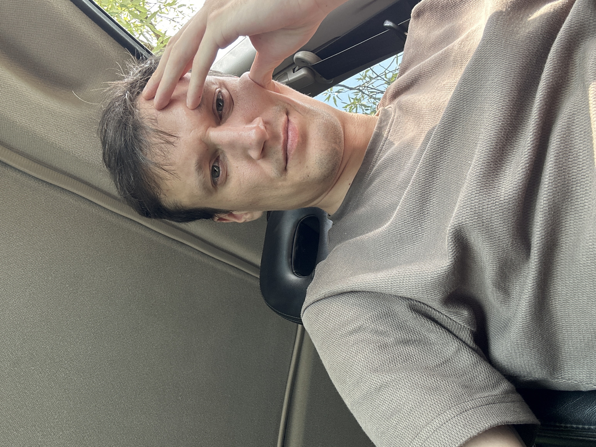 A man is sitting in the front seat of a car, with his hand resting on his forehead. The car interior is visible, including the headrest and part of the ceiling. Sunlight streams in from the side window, illuminating the man's face and clothing. The man is wearing a light brown t-shirt. The background outside the window shows greenery, suggesting an outdoor location. There is no information to ascertain the specific location within the city of Makiv, Ukraine.