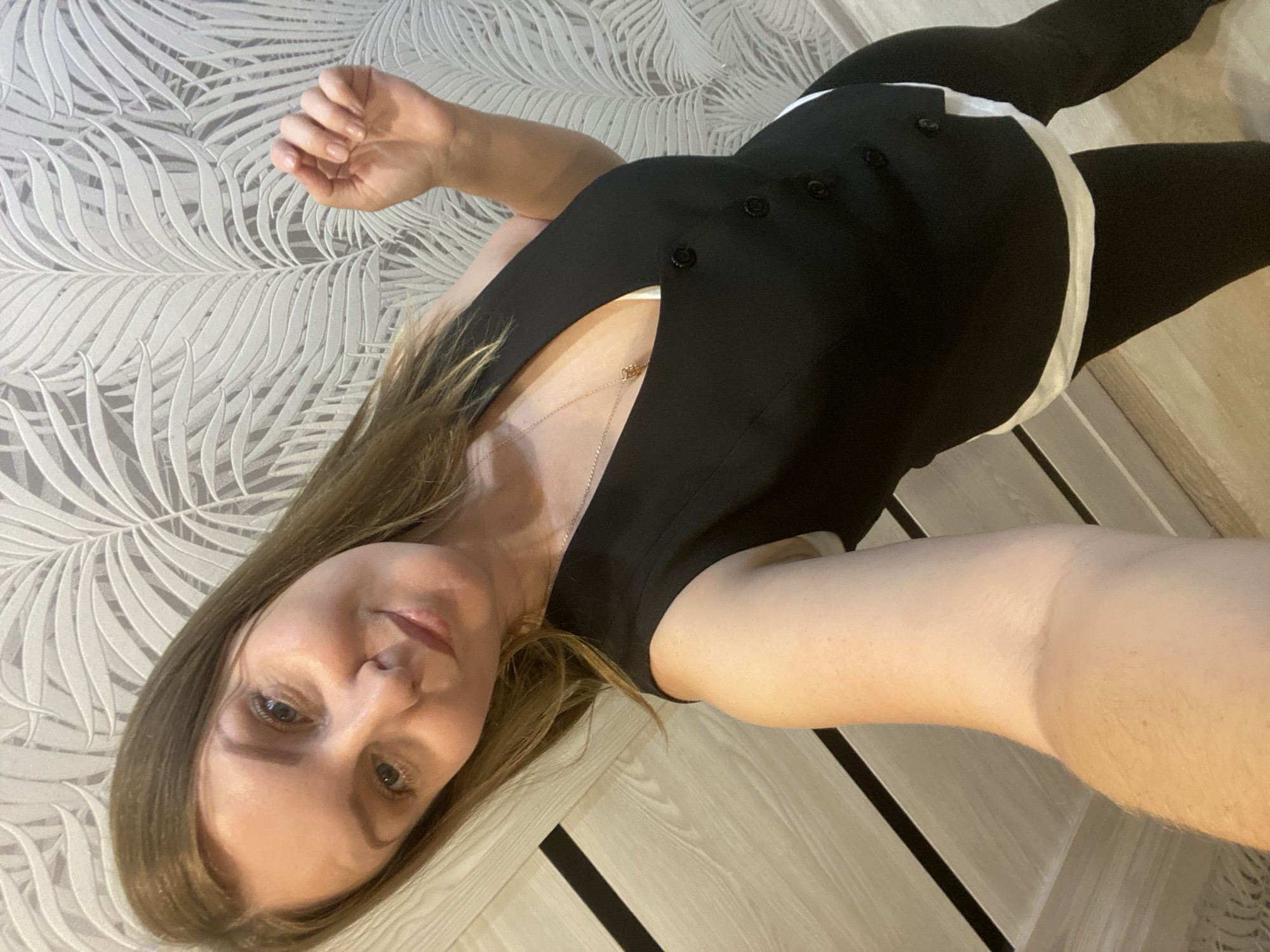 A young woman with shoulder-length brown hair takes a selfie in a room. She is wearing a black sleeveless vest over a white top, and black leggings. The woman is standing, and her left arm is extended towards the camera, holding the phone. In the background, there is a wall with a palm leaf patterned wallpaper and a wooden door. The floor appears to be light-colored wood. There is no indication of the location being the city of Noviy, Russia.