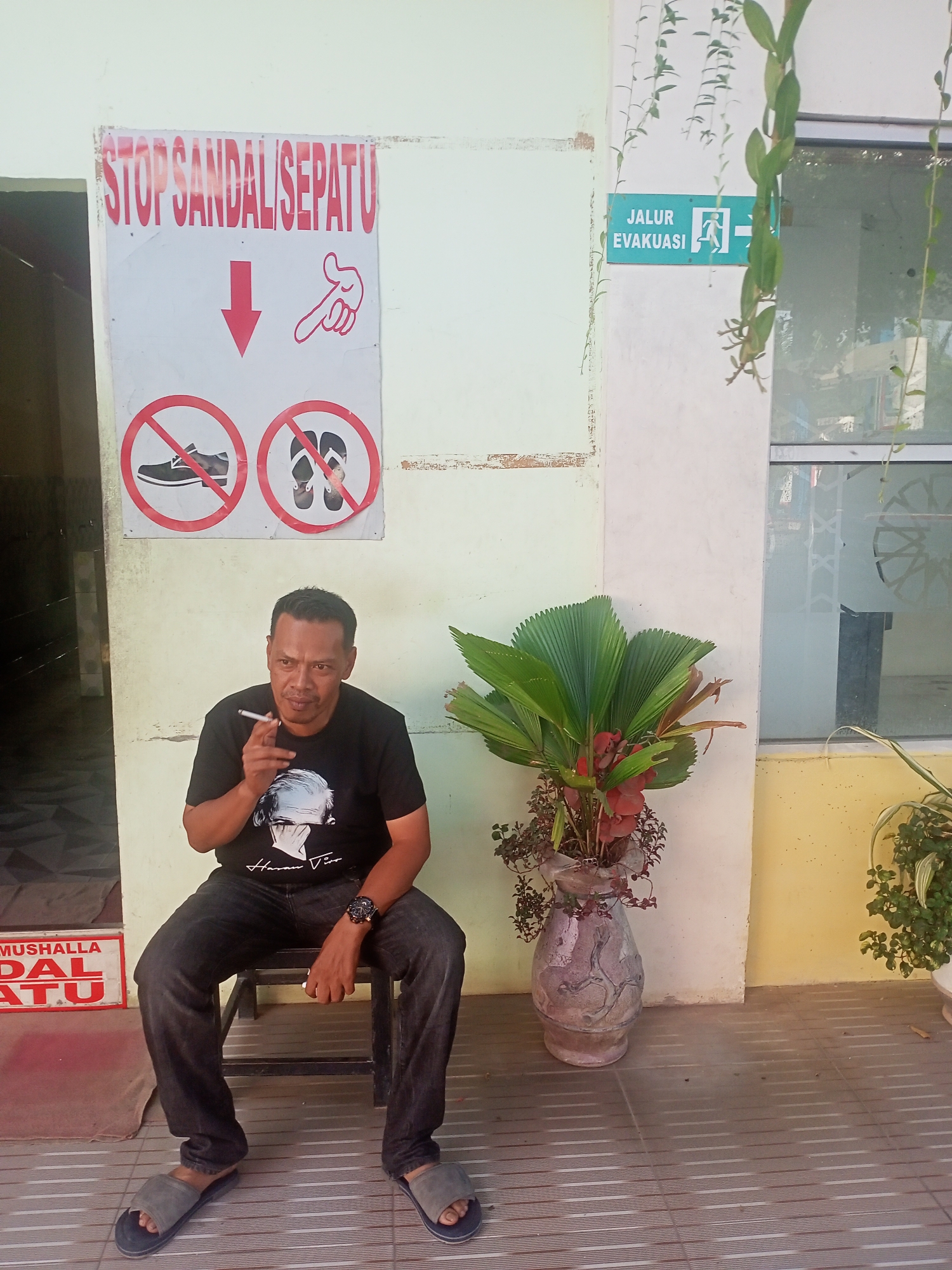 A man is seated on a low, dark stool, smoking a cigarette, in what appears to be a semi-outdoor or sheltered area of a building in Lhokdagang, Indonesia. He is of middle age with short dark hair, wearing a black t-shirt featuring a graphic of a person's face (possibly Albert Einstein) and script text, dark jeans, and grey sandals. A watch is visible on his left wrist.
To his right, a large fan-leafed plant with reddish accents is potted in a decorative, textured vase. The background consists of light green and yellow walls, a tiled floor with horizontal lines, and a window with some reflective patterns.
Several signs are visible. Above the man, a prominent white sign with red text reads "STOP SANDAL/SEPATU" (Stop Sandals/Shoes), accompanied by a downward arrow and a pointing hand symbol. Below this text, two red circular no-entry signs depict a shoe and a pair of sandals, reinforcing the prohibition of footwear. Further right, on a green background, a sign indicates "JALUR EVAKUASI" (Evacuation Route) with a white running man symbol and an arrow pointing to the right. Partially visible near the floor on the left is a red sign with white text that appears to say "MUSHALLA DAL ATU," likely indicating a prayer room where footwear is not permitted. The scene is well-lit, suggesting daytime.