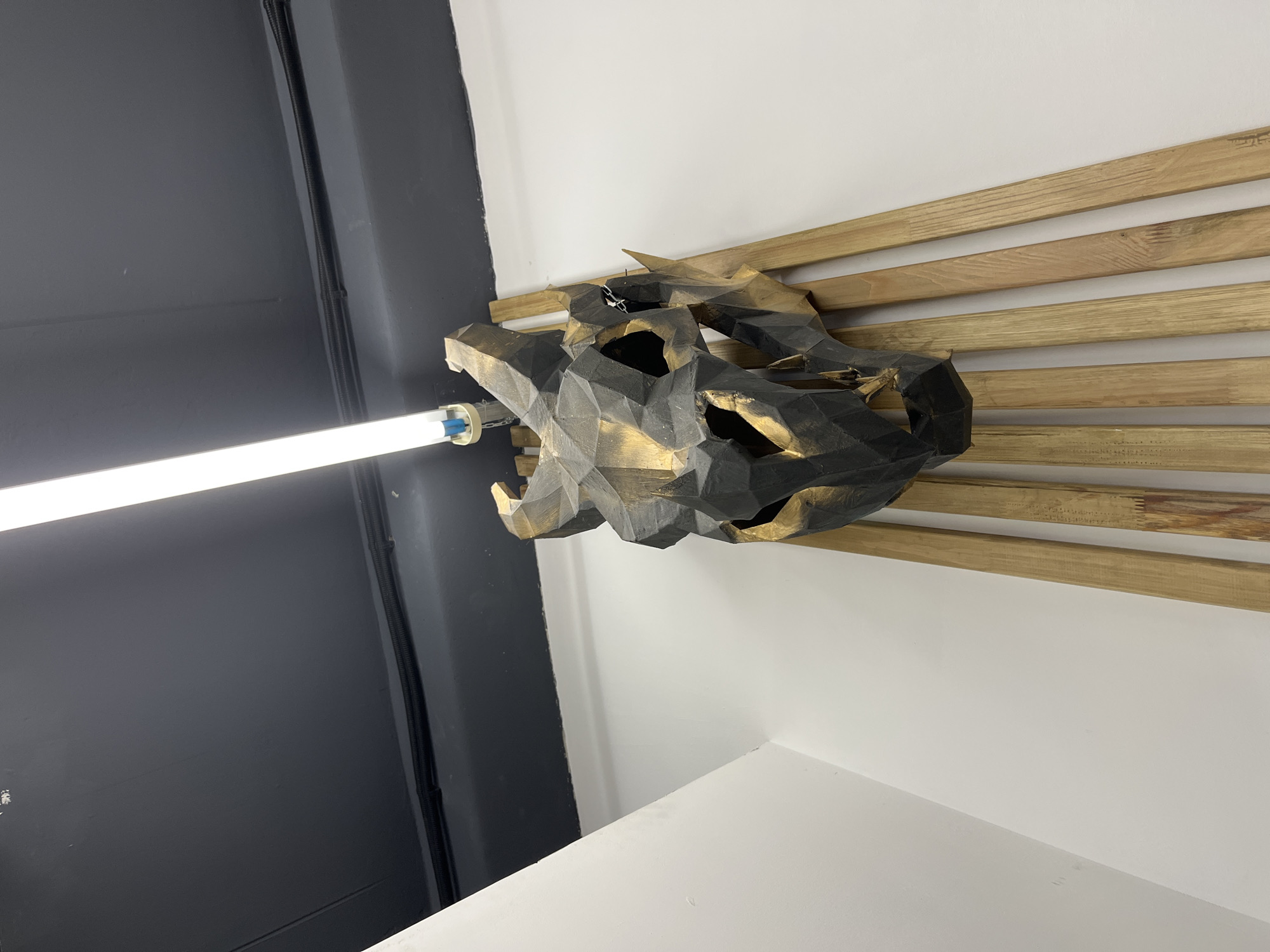An interior shot displays a striking geometric sculpture of a stylized animal head or skull, presented in dark tones with metallic gold accents. The multifaceted sculpture is mounted against a wall segment featuring several horizontal wooden planks of light, natural wood color.
To the left of the sculpture, a bright, horizontally oriented fluorescent tube or LED light bar is affixed to a dark grey wall, emitting strong white light. A dark cable or conduit runs vertically along this dark wall. The lower and right portions of the image reveal clean, plain white walls, suggesting a modern and minimalist design aesthetic. The distinct corner where the dark grey wall meets a white wall is visible.
The scene focuses solely on the art installation within its architectural environment, with no people or explicit activity taking place. The artificial lighting and indoor setting do not provide cues for the time of day or weather. No visible text is present in the image. This contemporary display is situated in Chaikovsky, Russia.