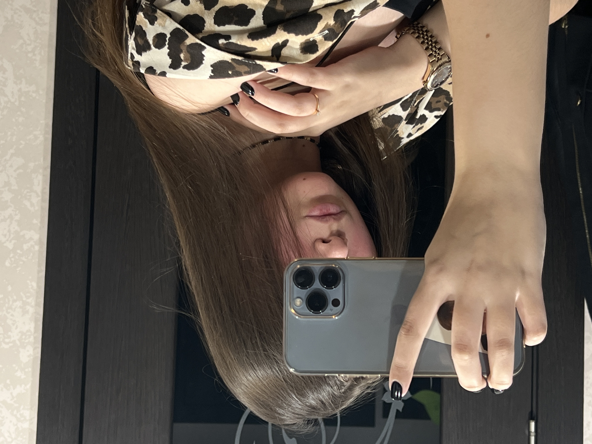 A woman with long brown hair, wearing an animal print top and a gold watch, takes a selfie with a gray iPhone. She has black nail polish and wears a ring on her left hand. The background includes a dark brown door and a patterned wall. The location is Chaikovsky, Russia.