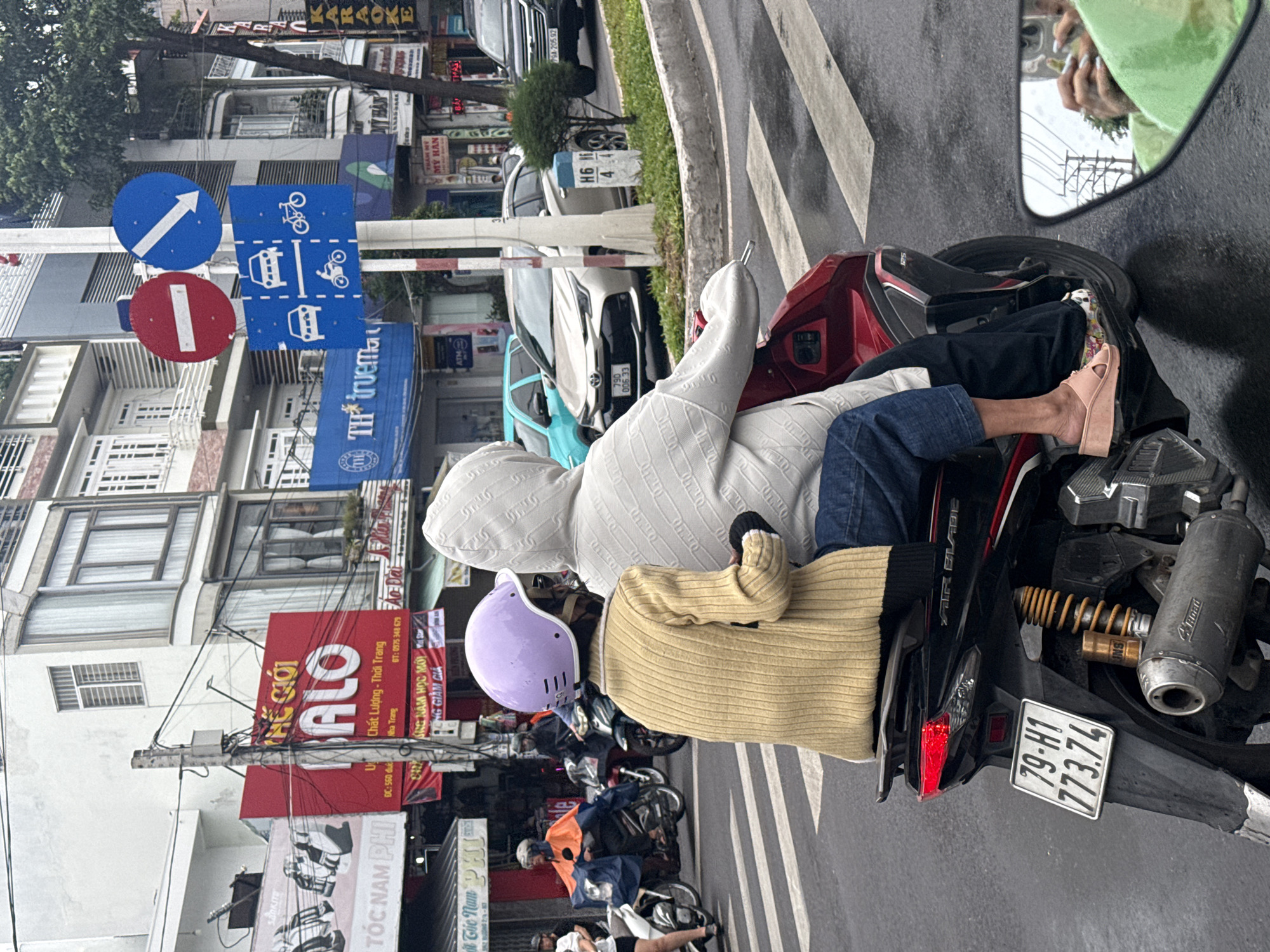 The image depicts a street scene in Nha Trang, Vietnam, with a focus on a motorcycle carrying two individuals. The rider, wearing a light brown sweater and a purple helmet, is in the foreground. Behind them, a passenger in a white hooded jacket is visible. Both are seated on a red motorcycle. The license plate of the motorcycle is clearly visible and reads "79-H1 773.74".
The environment shows a city street with buildings lining the background. Several of these buildings have signs with Vietnamese text, and one prominent red sign displays "ALO" in large white letters. Traffic signs are also present, including a red "no entry" sign and a blue sign with directional arrows and icons indicating vehicles and bicycles. Other vehicles, including cars and motorcycles, are visible in the background, suggesting normal traffic conditions. The road surface appears wet, indicating recent rain. In the lower right corner, a motorcycle's rearview mirror reflects the person taking the photo, holding a smartphone. The overall scene captures a moment of daily life in a Vietnamese urban setting.