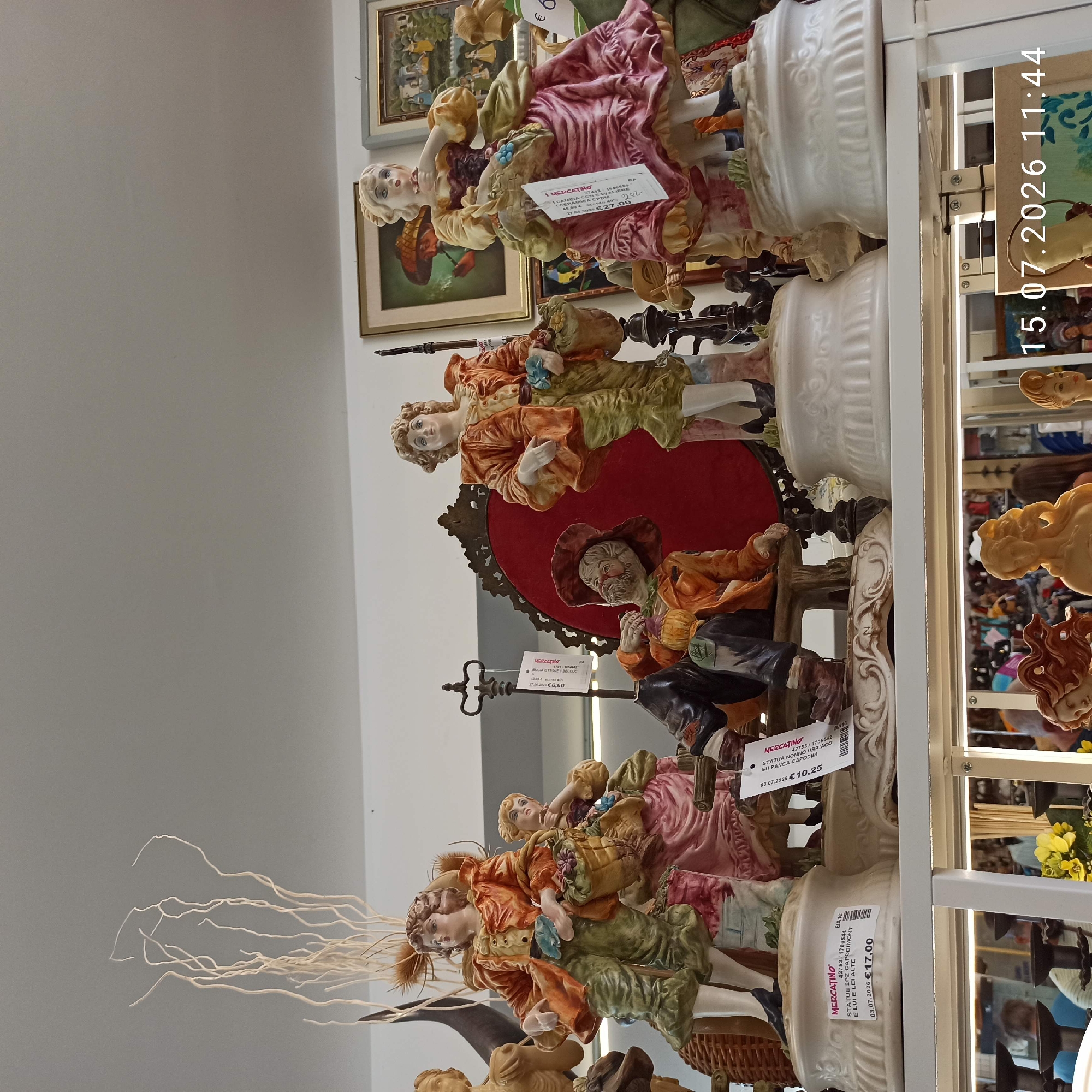 This image shows a display of ceramic figurines on a shelf, likely in a shop. The figurines depict people in historical or theatrical attire, carrying baskets of flowers and fruit. Several of the figurines are mounted on ornate white bases. Price tags are visible on some of the figurines, with dates and prices such as "03.07.2026 €17,00" and "03.07.2026 €10,25". In the background, framed paintings are visible on the wall, one depicting a man smoking a pipe and another with a landscape scene. The lighting suggests an indoor environment, possibly a shop or market. The bottom portion of the image reveals a shelf below with more items for sale, including what appear to be busts of women, and a timestamp overlaid on the image reads "15.07.2026 11:44". The question of the location in Molfetta, Italy, is not directly addressed by visual cues in the image.