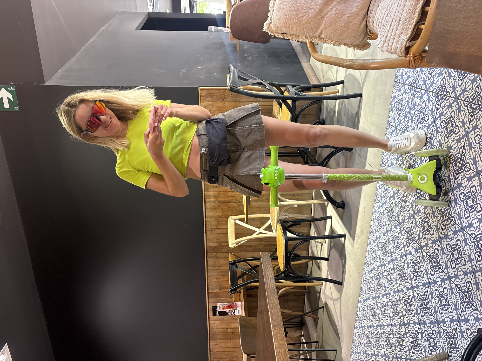 In this vertical, eye-level shot, a woman with blonde hair and bright orange sunglasses poses playfully indoors, holding a smartphone in her hands. She is wearing a neon yellow t-shirt and a khaki skirt with a black fanny pack. Her legs are extended, and her feet are clad in white sneakers, one of which is planted on the handle of a green scooter with a black deck. The scooter is positioned on a floor with a blue and white patterned tile design.
The background reveals a cafe or restaurant setting. To the left, a dark gray wall contrasts with a smaller section of a light gray ceiling, where a green directional arrow sign with white text is visible. In the middle ground, several chairs and tables are arranged, with some chairs featuring wooden frames and seats, while others are black metal with dark seats. A tall, light-colored chair with a woven cushion is visible in the upper right corner. The overall lighting suggests daytime, possibly late morning or afternoon, with some natural light filtering in. The woman's posture and smile convey a lighthearted and joyful emotion. The visible text on the arrow sign includes "LOYCO" and a stylized arrow.