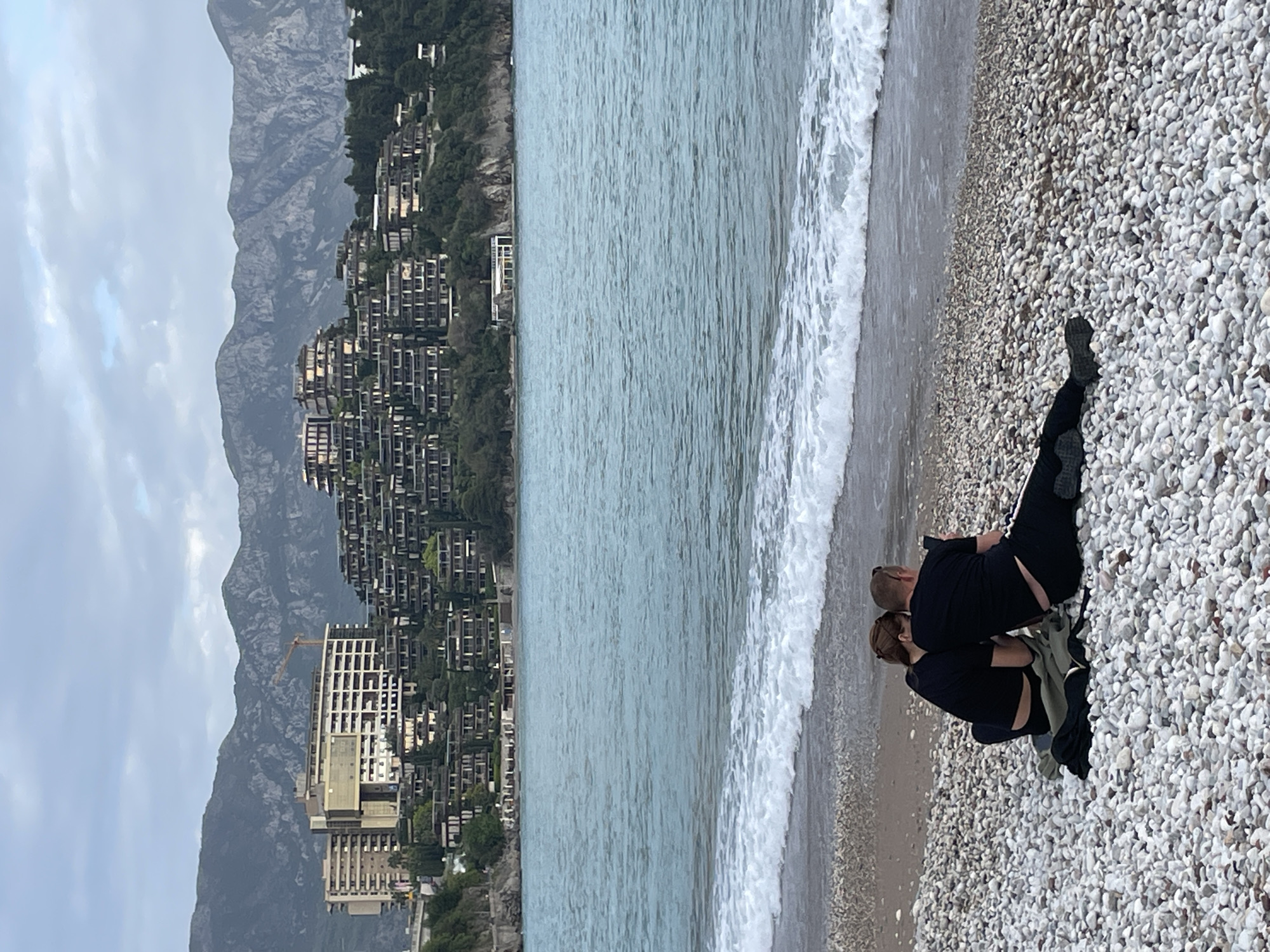 The image depicts a beach scene in Budva, Montenegro. The beach is composed of small white pebbles. A man and a woman are crouched down on the beach, seemingly interacting or looking at something. Behind them, the sea stretches out to the left, with waves breaking on the shore. In the background, a hillside is visible, covered with buildings, and further back, the mountainous terrain. The sky is overcast, and a construction crane can be seen among the buildings.