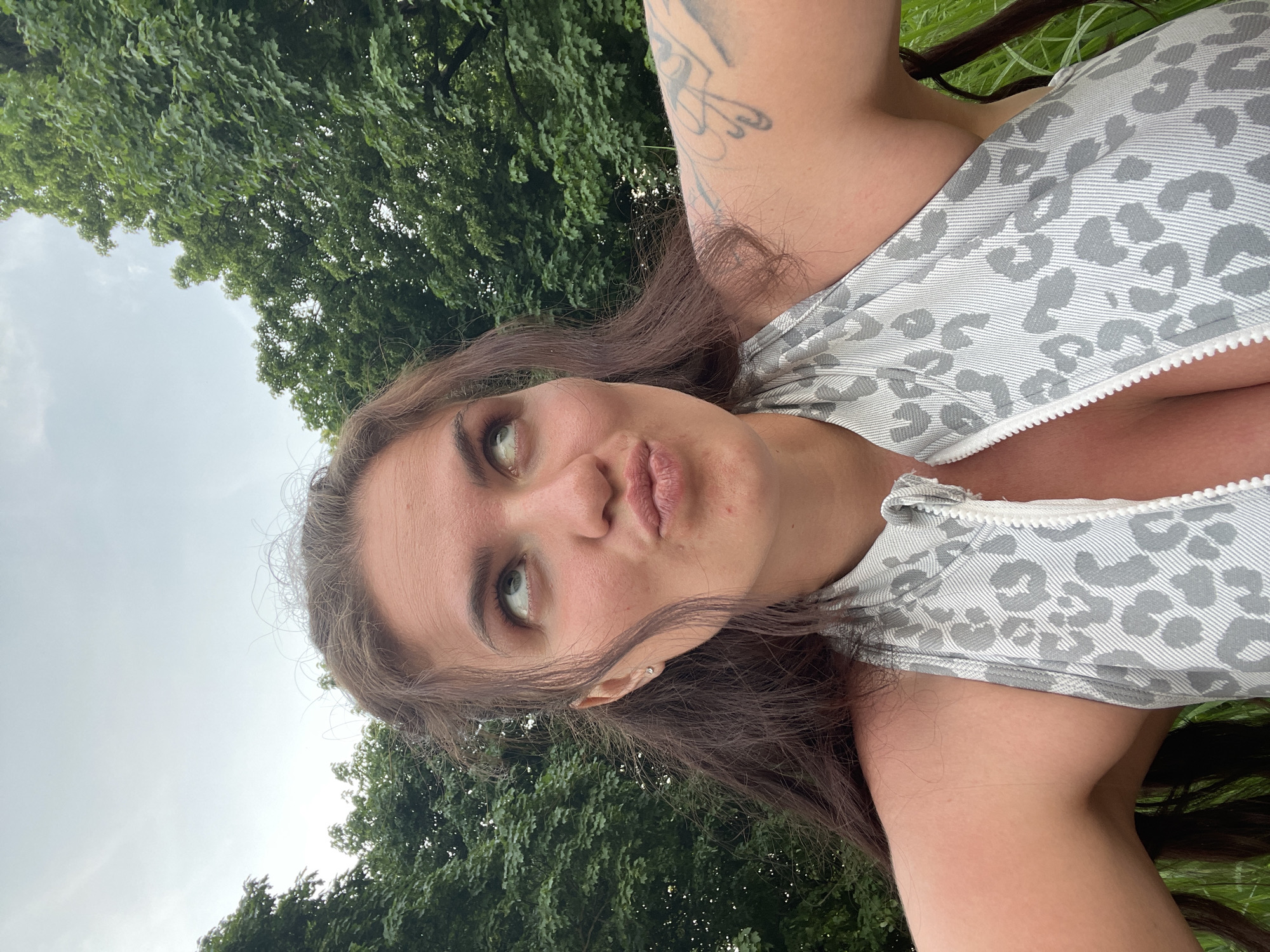 A woman is the primary subject, likely outdoors based on the presence of green foliage resembling trees in the background and the presence of the sky. She is wearing a gray and white patterned top with a zipper. She has long brown hair and is positioned at an angle relative to the camera. A tattoo is visible on her upper arm. The location in Ostrov, Russia, cannot be verified.