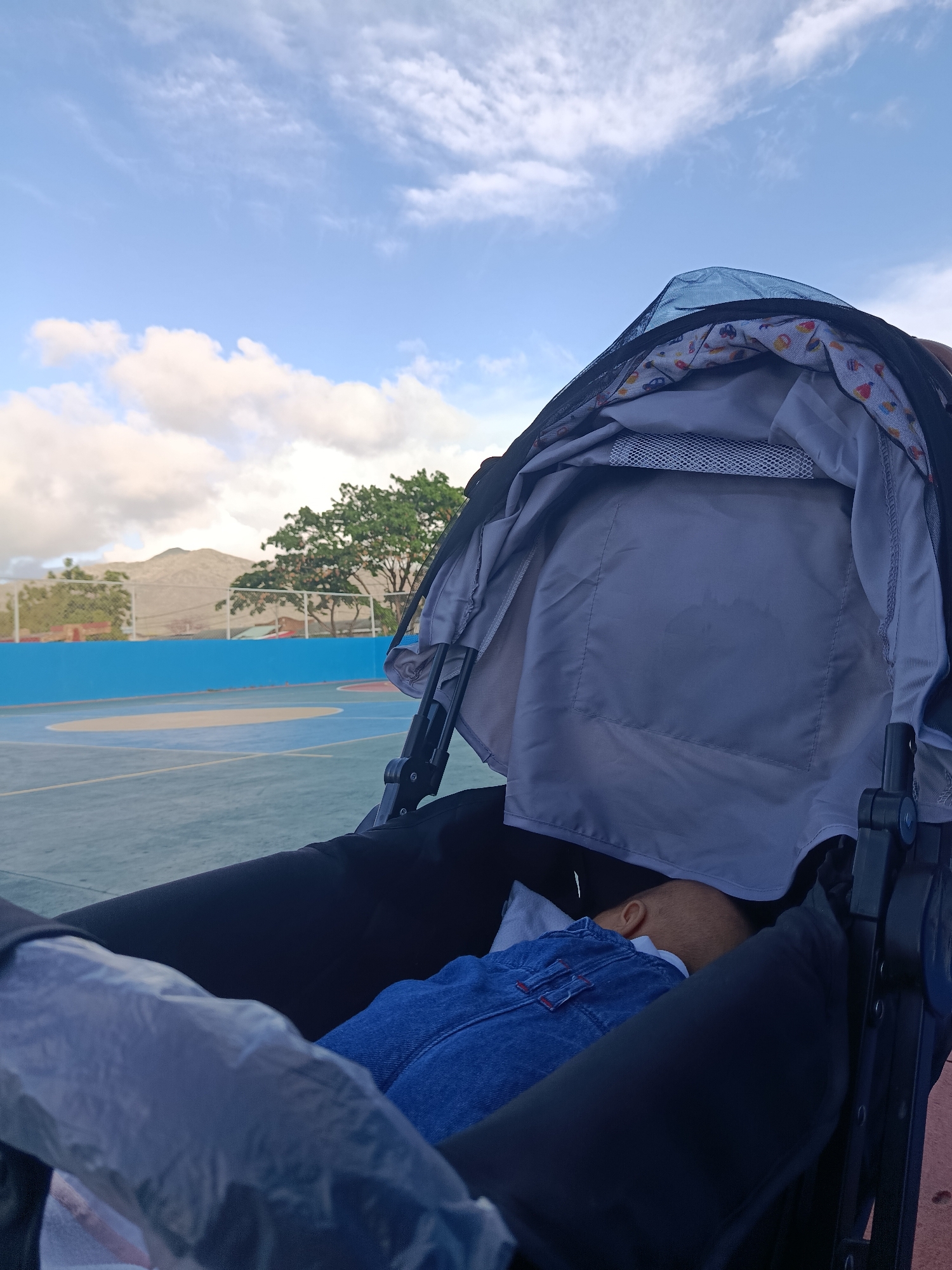 An infant sleeps in a stroller parked on an outdoor basketball court under a partly cloudy sky. The stroller is black with a grey canopy, and the infant is dressed in blue denim overalls. In the background, a blue-lined basketball court is visible, with a white fence and green trees. Mountains rise in the distance. The image appears to have been taken in Juan Griego, Venezuela.