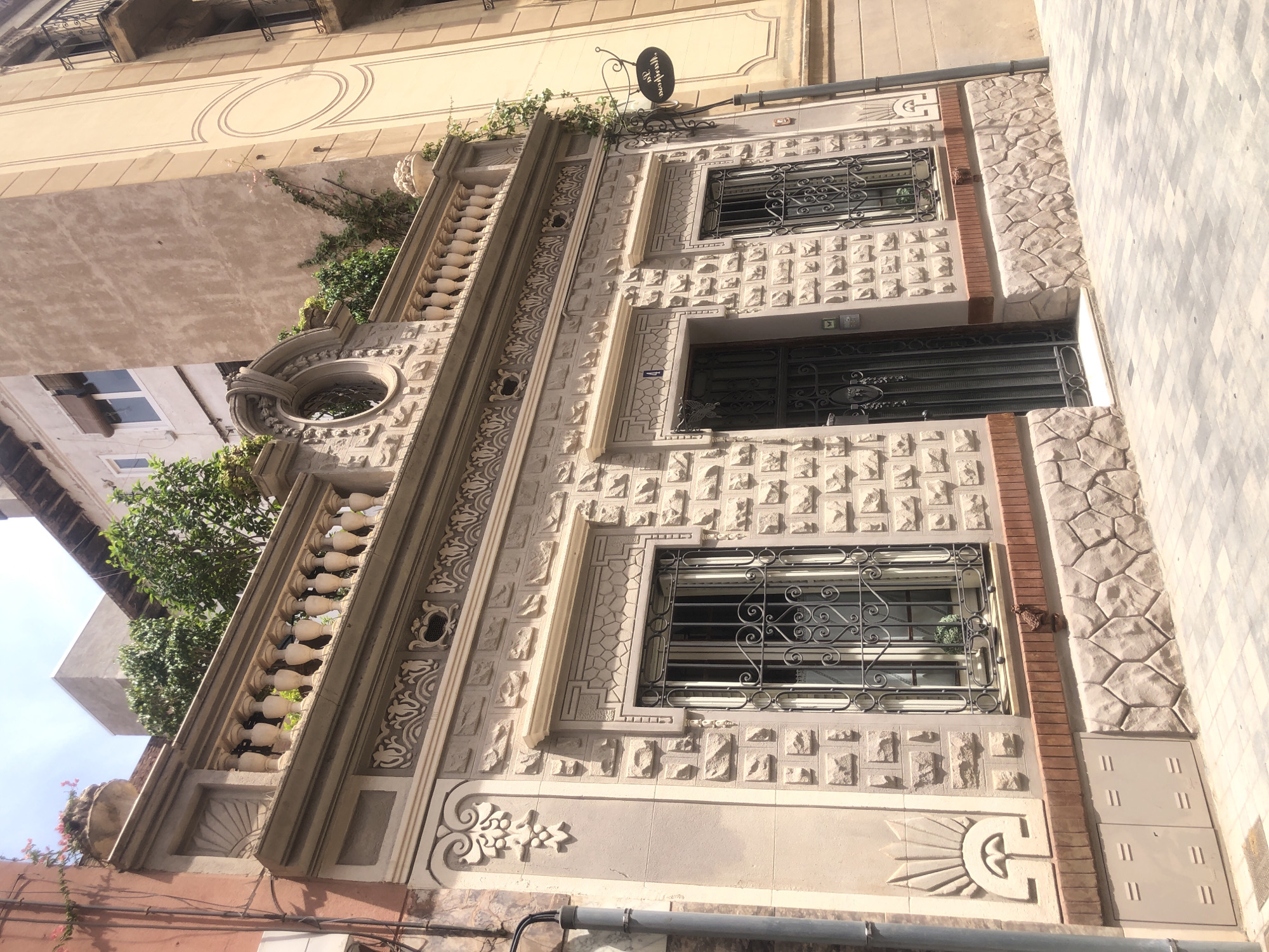 The image shows the exterior of a two-story building with intricate architectural details, located on a sunny day in a city. The building's facade is constructed with light-colored stone blocks, with decorative elements such as carved reliefs, ornamental moldings, and a wrought-iron balcony. There are two windows on the ground floor with protective iron grilles and a metal gate. A sign hanging from a pole on the right side of the building is partially visible. The ground in front of the building is paved with square tiles. The upper part of the building is partially obscured by a taller, adjacent building. Lush greenery is visible on the balcony of the lower building and also on the adjacent building. The sky is clear and blue.