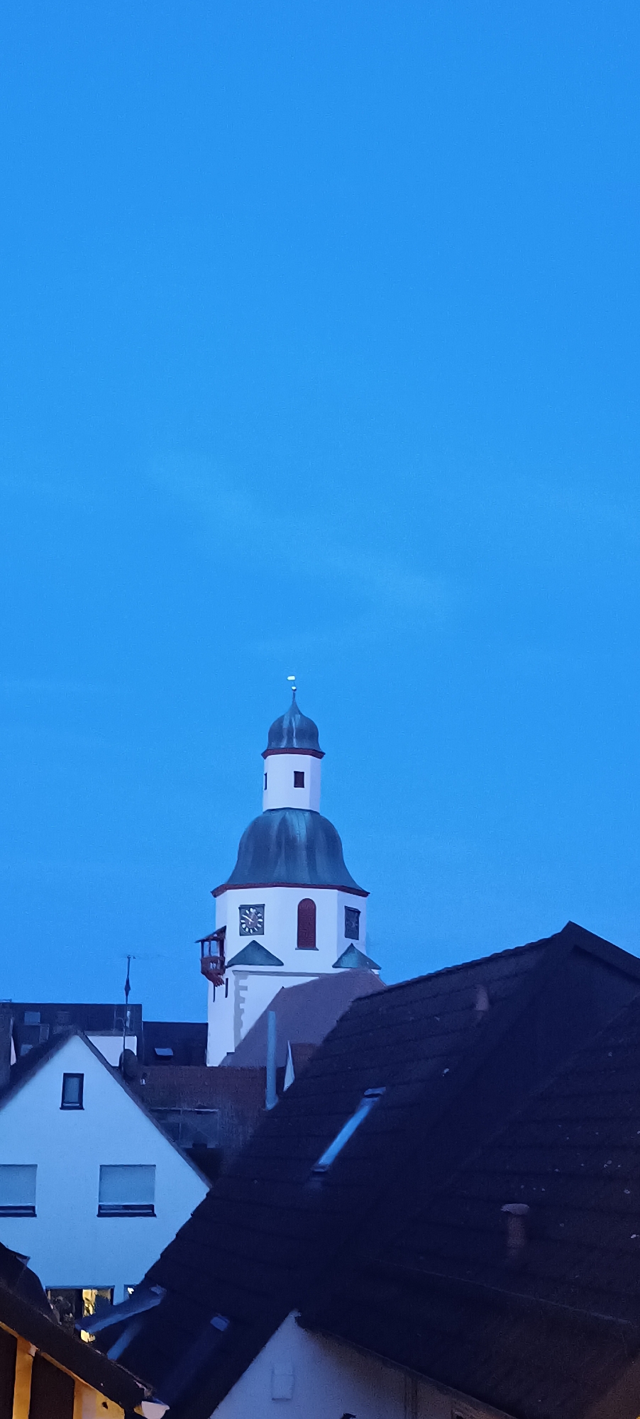 The image shows a view of the town hall tower in Winnenden, Germany, against a clear blue sky. The tower is tall and white, with a dark green, domed roof. It has a clock on one side and several windows. In the foreground, there are rooftops of residential buildings, some with dark shingles and others with white facades. The lighting suggests it is either early morning or late evening. There are no people visible in the image.
