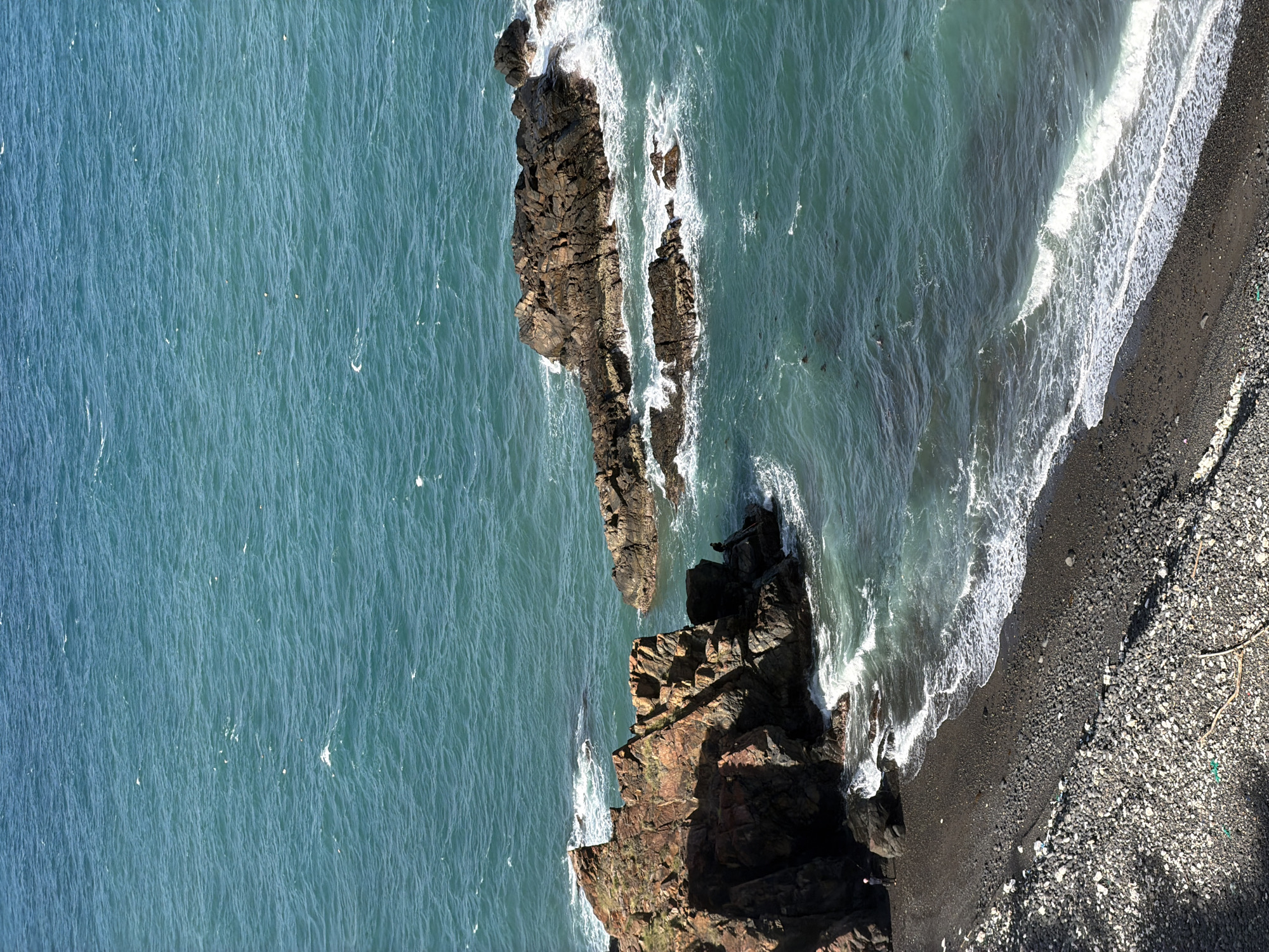 An aerial view depicts a rugged coastline with rocky cliffs and a dark-sand beach. The ocean, with its turbulent waves crashing against the rocks and shore, occupies the majority of the frame. The cliffs are a mix of dark brown and gray, appearing weathered and jagged. The beach consists of small, dark pebbles. The location is in Nam Nha Trang Ward, Vietnam. No people are visible in the image.
