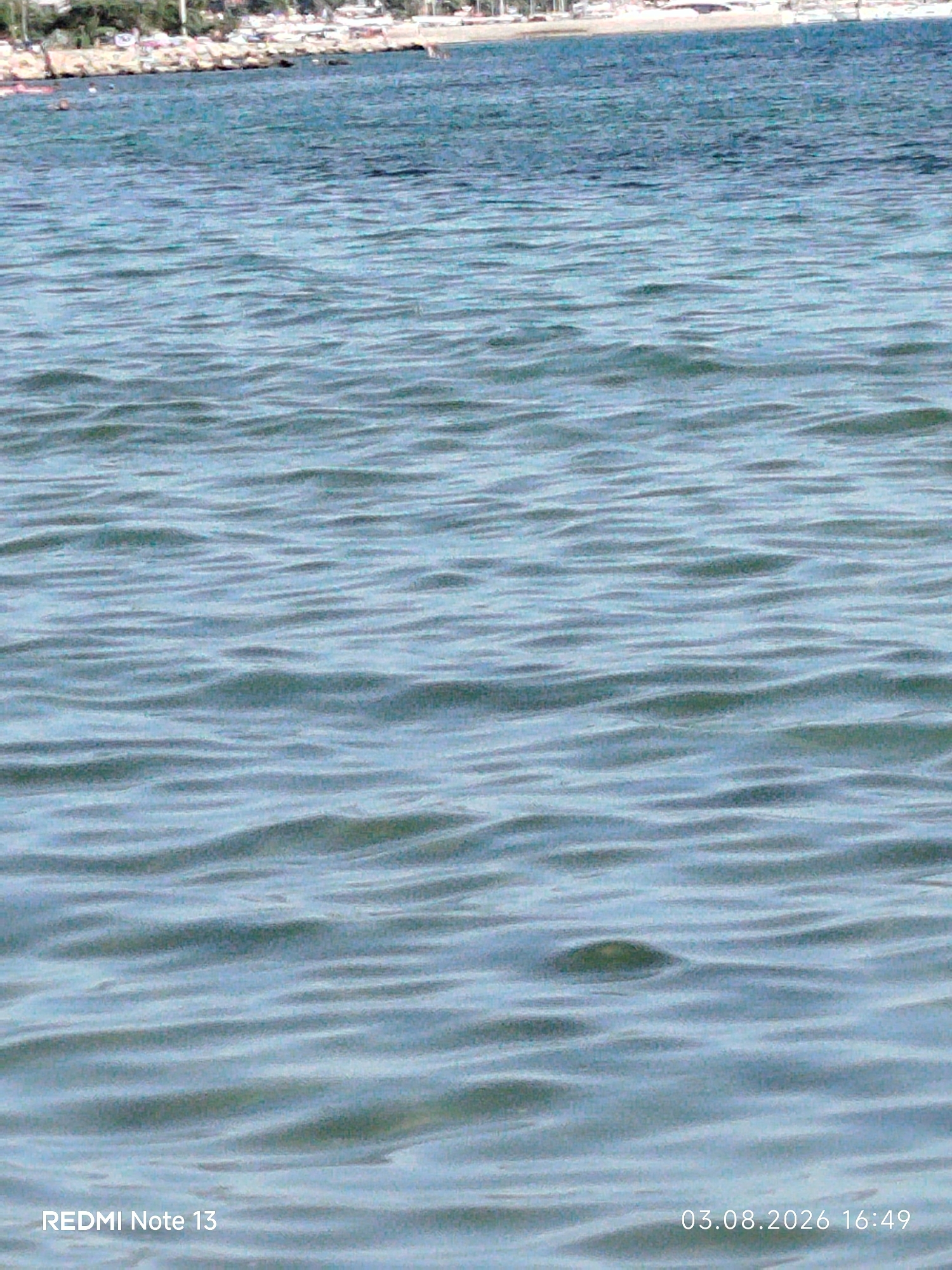 The image displays a body of water with gentle ripples, extending from the foreground to a blurred background. In the distant background, a shoreline is visible, characterized by a strip of land with buildings and trees. The water itself is a shade of blue-green, with variations in light and shadow creating a textured appearance. The lower portion of the image contains text overlaying the water, indicating "REDMI Note 13" on the left and "03.08.2026 16:49" on the right. The scene suggests an outdoor, coastal environment, likely a beach or harbor area within the Sithonia Municipal Unit, Greece, given the textual watermark. There are no discernible people or specific actions captured in the frame, focusing solely on the natural element of the water and the distant, indistinct shoreline.