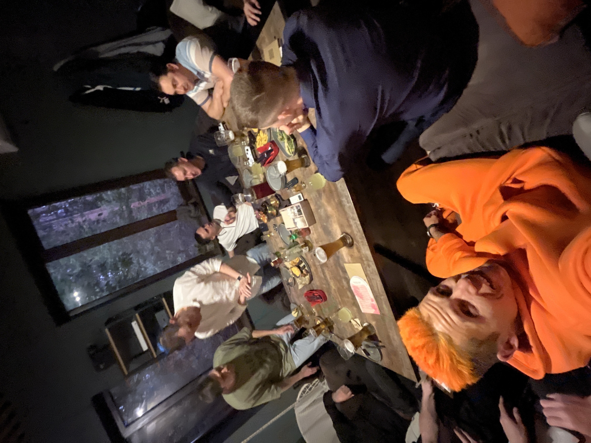 The image captures a casual social gathering of a group of approximately seven to eight men around a long, rustic wooden table. The setting appears to be an indoor space, likely a bar or restaurant, illuminated by dim lighting, suggesting it is evening or night. The composition is tilted, providing an informal, overhead view of the scene.
The men are dressed in casual attire, including t-shirts, long-sleeved shirts, and hoodies. Many are engaged in conversation, looking at each other or towards the center of the table. A man in a bright orange hoodie with distinct orange and blonde hair is prominently featured in the foreground, smiling or grinning directly at the camera.
The wooden table is laden with various beverages and food items. Multiple glasses of beer and cocktails, along with bottles and other drinkware, are scattered across its surface. Plates containing food, including French fries and a green salad, are also visible. Personal items such as mobile phones and what appears to be a menu or a piece of paper can also be seen. Large windows in the background show a blurry, dark outdoor scene, indicative of nighttime, with hints of trees or foliage. The atmosphere seems relaxed and convivial. The location context is Chaikovsky, Russia. No legible text is visible within the image.