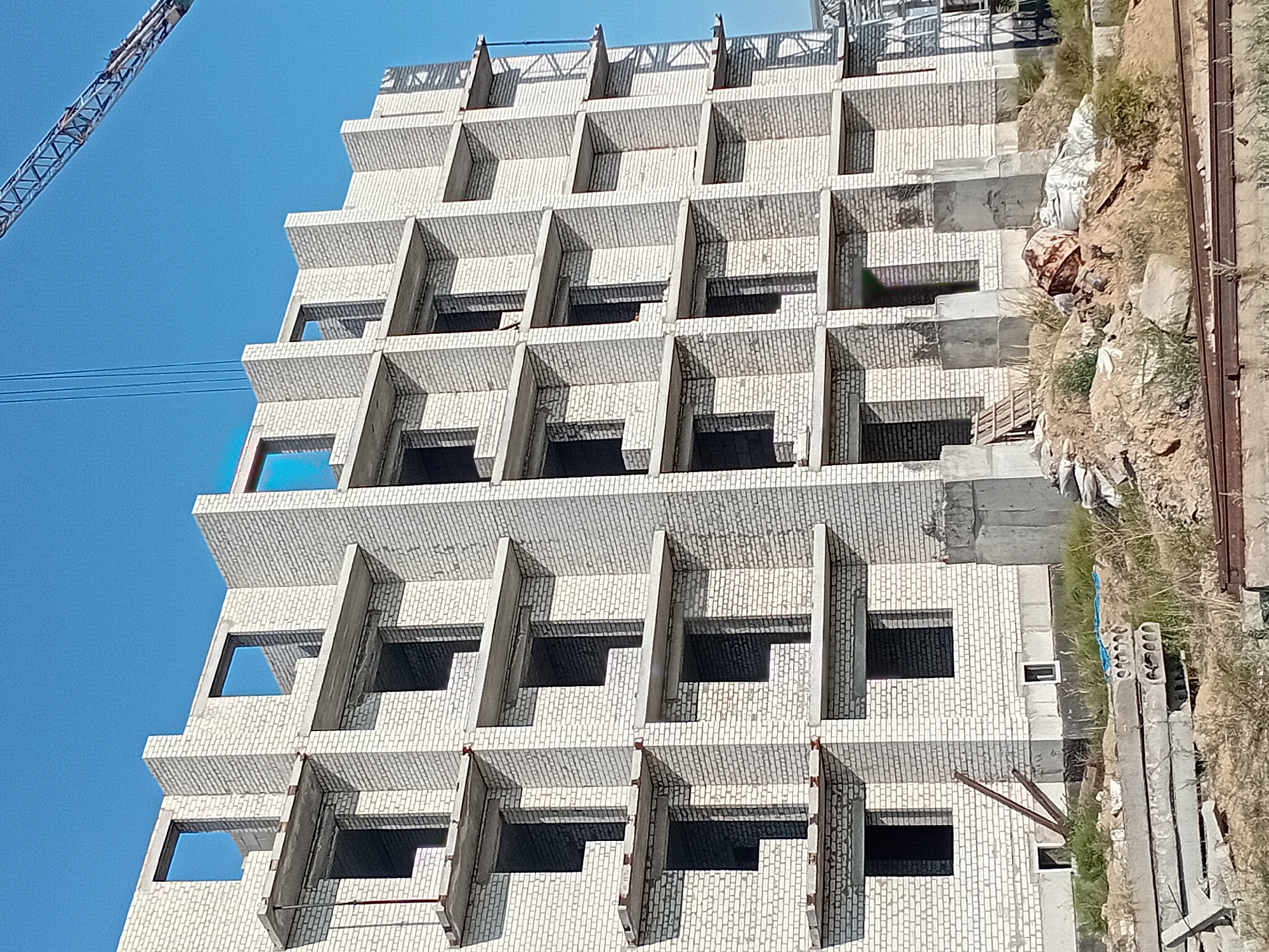The image depicts the construction of a multi-story building in Semey, Kazakhstan, under a clear blue sky. The building is constructed from white bricks and features numerous open window frames and concrete balconies. A construction crane is visible in the upper right portion of the image. The foreground shows a construction site with piles of dirt, debris, concrete slabs, and metal rebar. Vegetation is growing around the construction materials. The scene appears to be a typical day at a construction project.