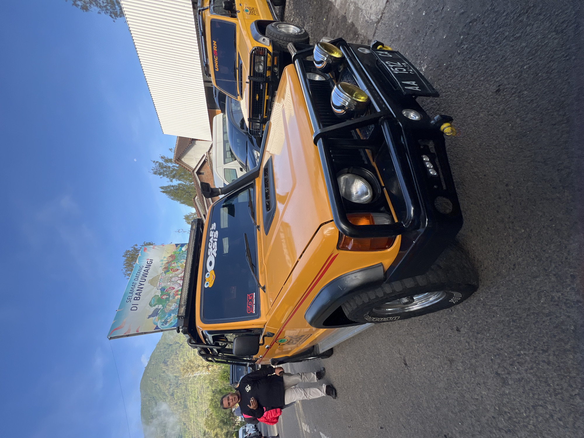 The image depicts two yellow Suzuki Jimny-style off-road vehicles parked on an asphalt road in an outdoor environment. A man stands beside the first vehicle. The front vehicle has a black bull bar with auxiliary lights and a license plate that reads "AA 1512 CA". The first vehicle has a snorkel, and an advertising banner is attached to its roof. The other vehicle in the background has a "UNICORN" logo on its windshield. The background includes a hillside covered with vegetation and a white building. The sky is blue. The location is near Banyuwangi, Indonesia, as indicated by the advertising banner on the first vehicle.