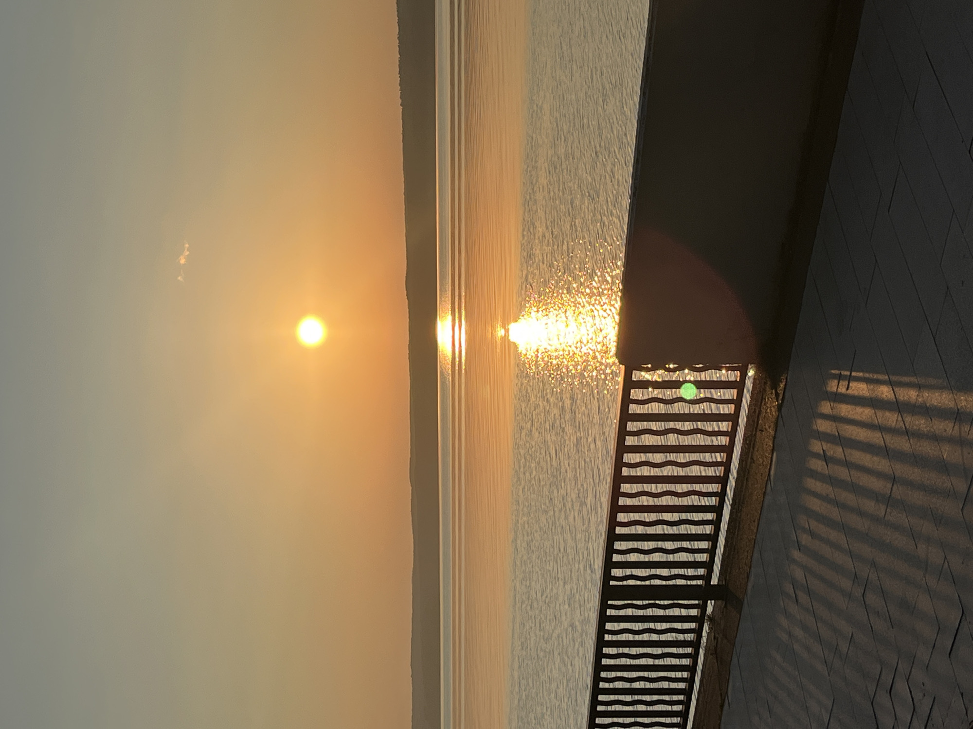 The image depicts a tranquil sunset over a wide body of water, likely the Kama River in Chaikovsky, Russia. The sun, a bright orange orb, is positioned in the upper central portion of the sky, casting a golden reflection on the calm water's surface. A hazy, distant shoreline with a gentle slope forms the horizon. In the foreground, a dark metal fence with vertical, wavy bars separates a paved walkway from the water. The walkway, paved with rectangular stones, displays elongated shadows cast by the fence and possibly other unseen structures, indicating the low angle of the setting sun. The overall scene is serene and bathed in the warm, diffused light of twilight. There are no people visible in the image.