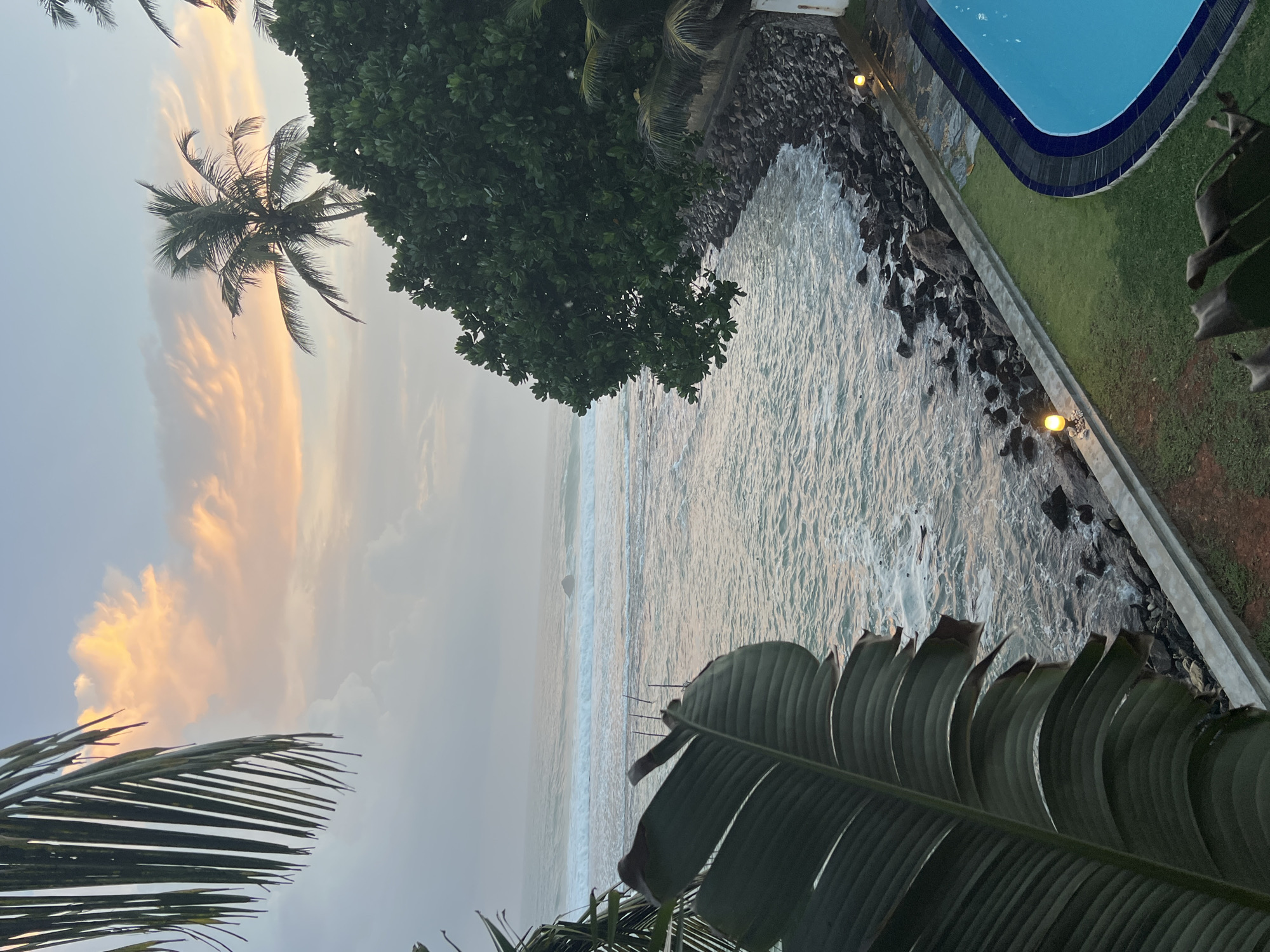 This is a medium-shot, eye-level, horizontal, outdoor, high-angle photograph of a coastal scene at dusk. The ocean fills the majority of the frame, with waves breaking on a rocky shoreline. In the foreground, a large, dark green leaf from a tropical plant partially obscures the view. To the right, a swimming pool with bright blue water is visible, bordered by a well-manicured lawn and a low wall. The shoreline is lined with dark, jagged rocks. Scattered along the wall are small, yellow outdoor lights. In the distance, palm trees and lush green foliage frame the scene. The sky above is a mix of soft grays and pastel pinks and oranges, indicating either sunrise or sunset. The overall mood is tranquil and serene. This image was taken in Goviyapana, Sri Lanka.