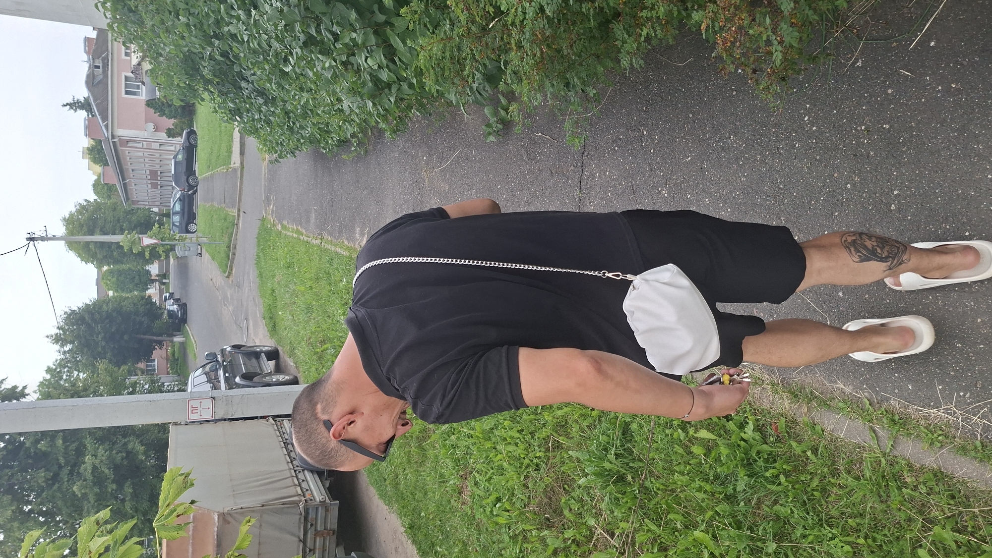 A man is walking away from the camera down a sidewalk next to a grassy area. He is wearing a black polo shirt, black shorts, and white slide sandals. He has sunglasses on and a white purse with a silver chain strap is slung across his body. He has a tattoo of a skull on his right calf. In the background, there are parked cars, trees, and buildings. The image appears to have been taken in Myadzyel, Belarus, a town known for its natural beauty and proximity to lakes.
