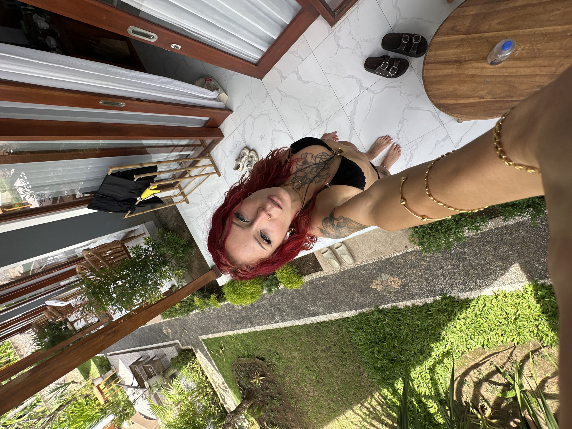 A downward, selfie-style perspective captures a red-haired woman in a black bikini on a tiled balcony. The woman is fair-skinned, with visible tattoos and a nostril piercing. Her left arm, adorned with gold bracelets, extends toward the camera.
The balcony overlooks a courtyard with a stone path flanked by lush greenery and tropical plants. Beyond the courtyard are structures, suggesting a villa or hotel environment.
The balcony includes wooden doors with white curtains, a wooden drying rack with garments, sandals, and a round wooden table with a small bottle on top.
Based on the architectural style and vegetation, the location is likely in a tropical region, consistent with Nusa Penida, Indonesia.