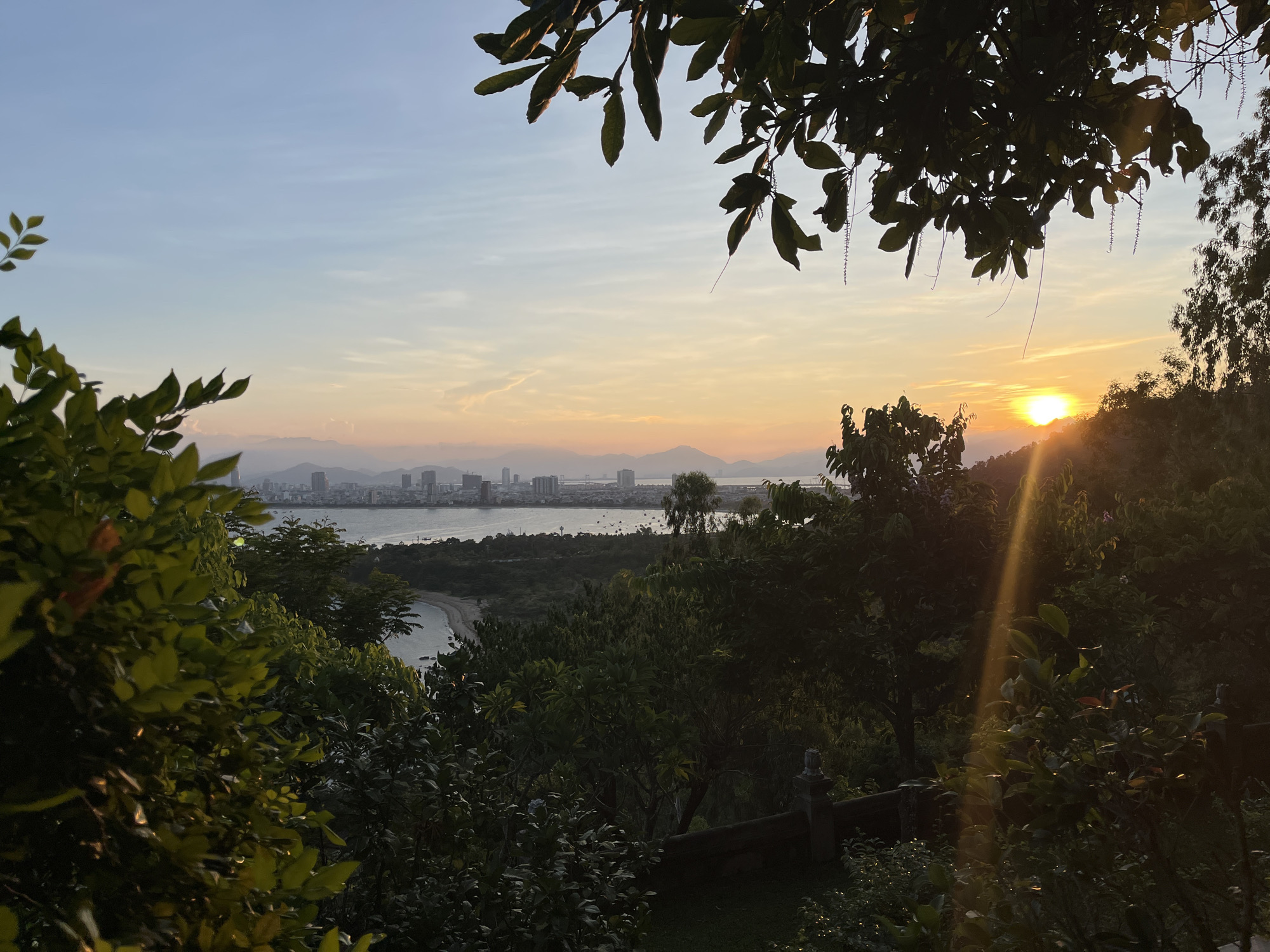 The image shows a view of Đà Nẵng, Vietnam, at sunset from a high vantage point surrounded by lush vegetation. In the distance, the skyline of Đà Nẵng is visible across the bay, with buildings and mountains faintly silhouetted against the sky. The sun is setting on the right side of the frame, casting a ray of light through the trees. The foreground is dominated by foliage, with branches and leaves framing the scene. A beach is visible in the middle distance, along with a small boat. The sky is a gradient of light blue and orange, indicative of sunset.