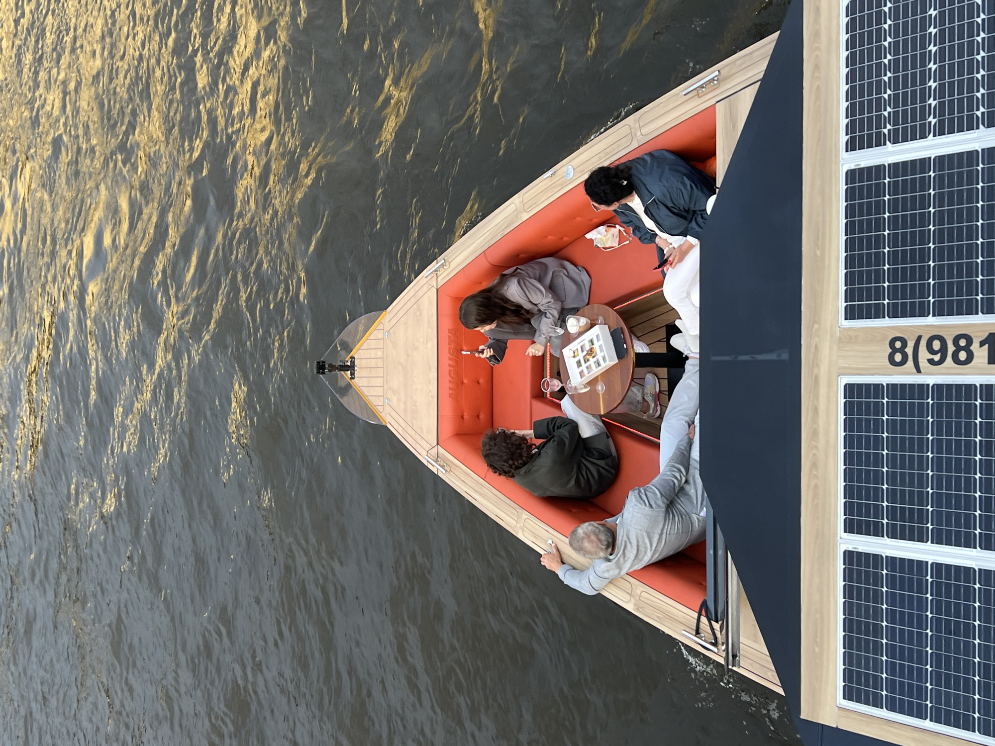 This aerial shot captures a group of four people enjoying a relaxing outing on a boat in Saint Petersburg, Russia. The boat features an orange upholstered seating area and a circular table. One person is seated on the left, looking at their phone, while another sits across from them, engaged with a drink. To the right, two more individuals are seated, one leaning back and the other looking towards the table. The boat is equipped with solar panels on its exterior, suggesting it is a solar-powered vessel. The water surrounding the boat appears calm with sunlight reflecting off its surface, indicating it's likely daytime, possibly late afternoon due to the warm light. The scene suggests a leisurely social gathering, perhaps a tour or a private outing on the city's waterways. Visible text on the boat reads "8(981".
