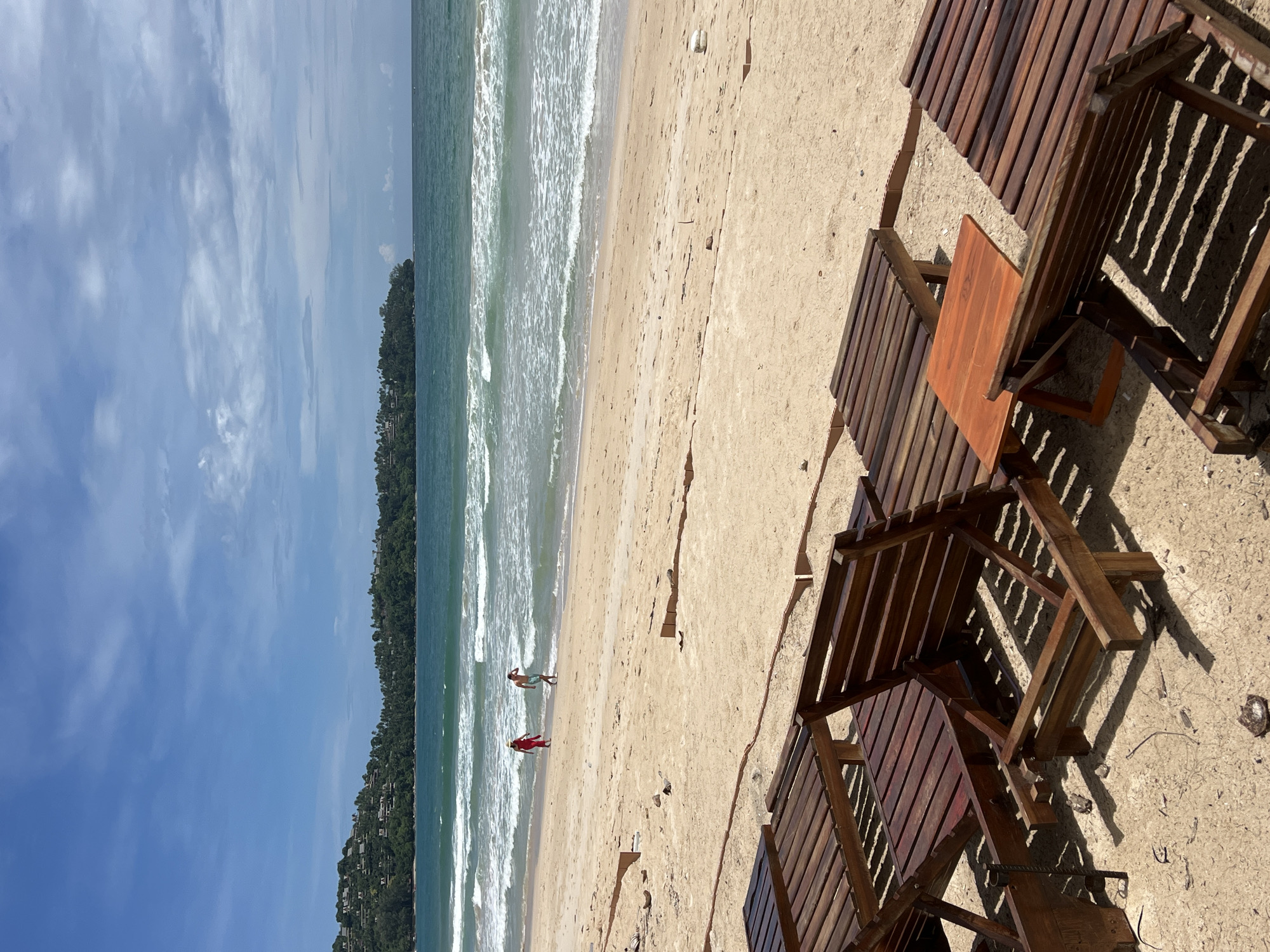 The image depicts a beach scene. In the foreground, several wooden lounge chairs are situated on the sand. In the middle ground, two people are standing in the water. One person is wearing a red outfit, while the other is bare-chested. The background shows a coastline with trees and buildings. The sky is blue with some clouds. The location is Unknown, Unknown.