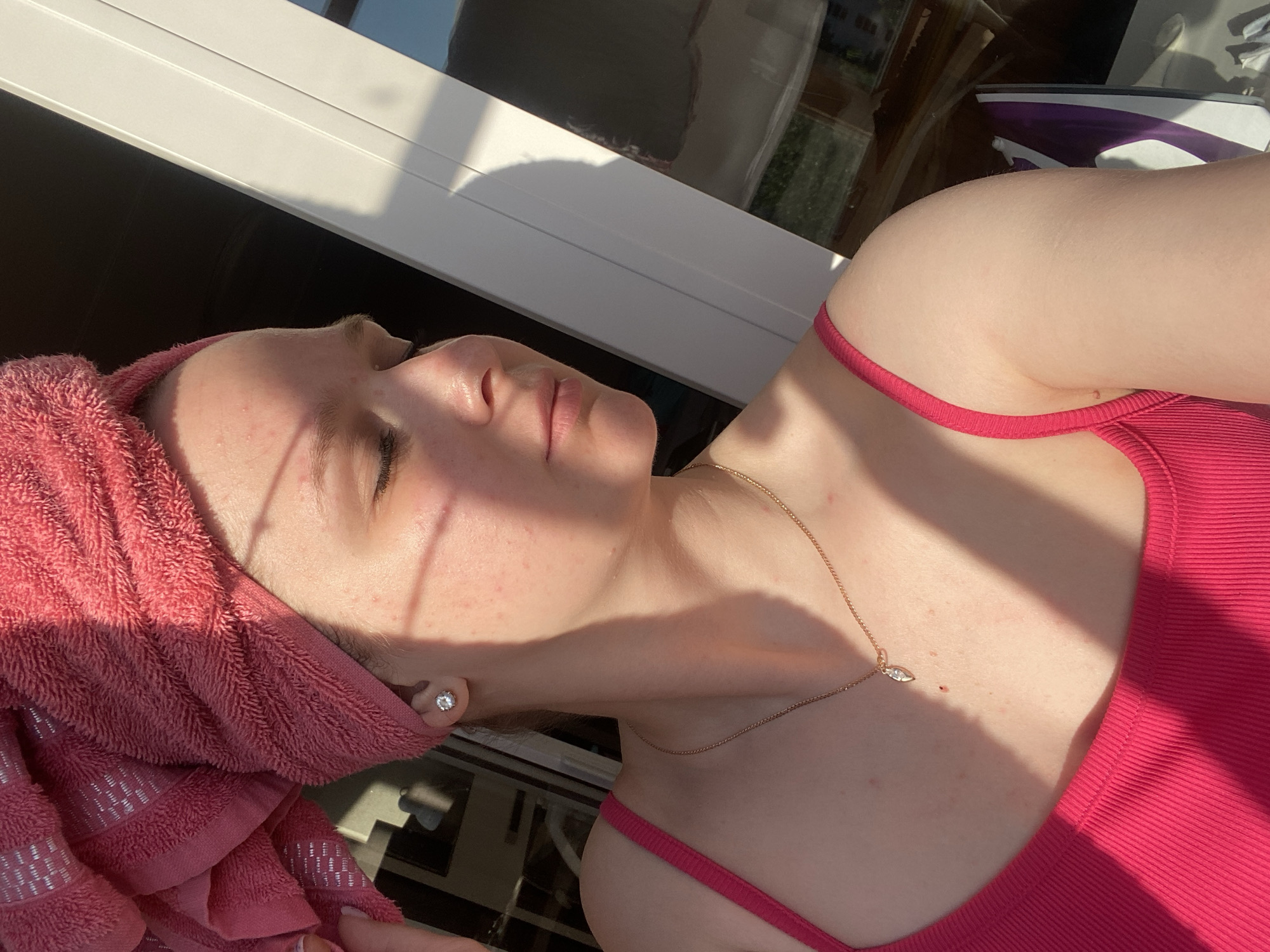 A woman is positioned in front of a window in an unidentified location within the city of Unknown, Unknown. She is wearing a pink tank top and a gold necklace with a pendant. Her hair is wrapped in a pink towel. An iron is visible in the background near the window. Sunlight is streaming in through the window, casting shadows on her face and upper body.