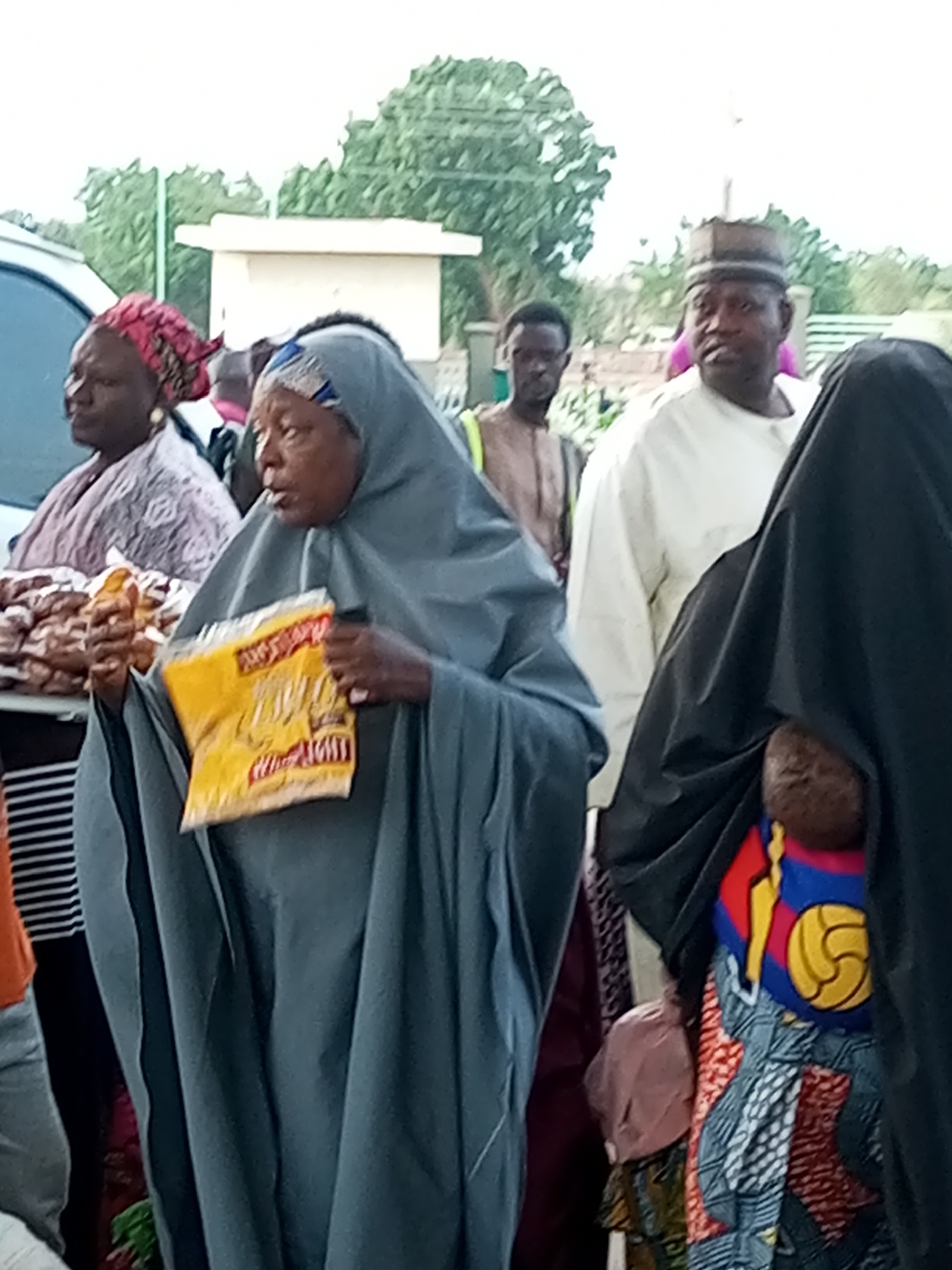 In Gombe, Nigeria, a group of people are gathered outdoors. The central figure, a woman, wears a grey hijab and holds a yellow food package. To her left, another woman is visible, wearing a patterned headscarf. Behind them, a man in a white garment and a cap stands, while another man in a brown robe is further back. A portion of a white vehicle is on the far left. The environment appears to be a market or a public gathering space, with a building and trees in the background.