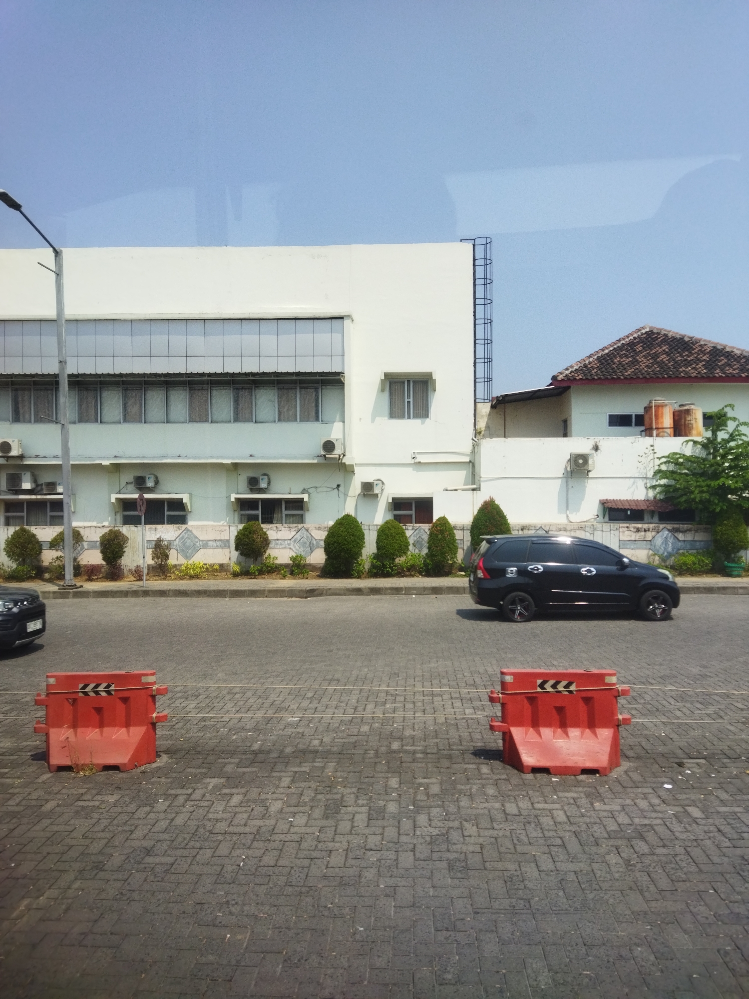 The image captures a street scene with a building and a black SUV. The building is white with a long row of windows. To the right of the main building, there is a smaller white building with a brown tiled roof and two cylindrical water tanks. In front of the buildings, there is a paved area with a black SUV driving on it. The foreground shows a cobblestone-like pavement with two bright orange traffic barriers. The sky is clear and blue. The overall setting appears to be an urban or industrial area in Grogol, Indonesia.