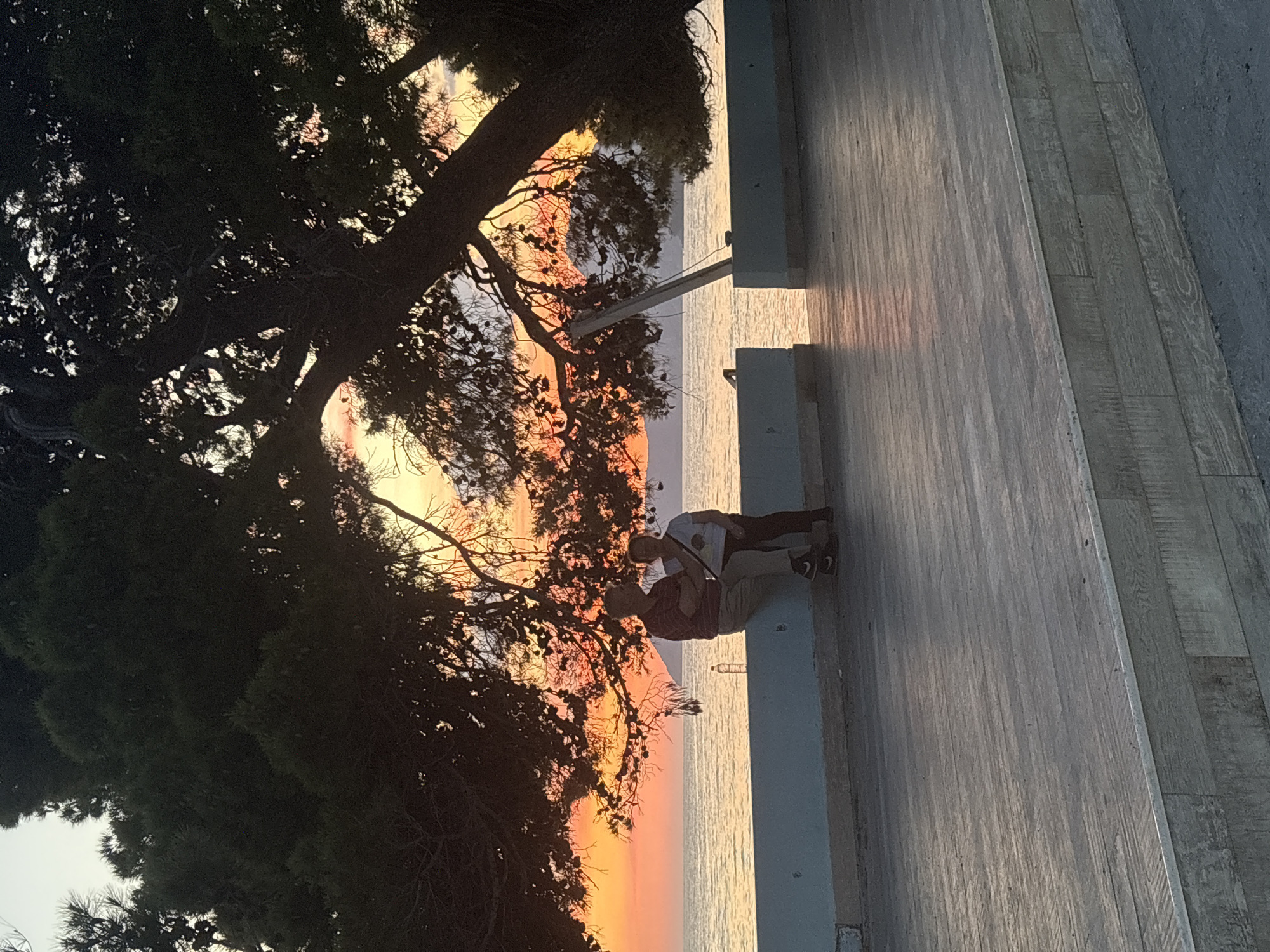 Captured at a horizontal angle, the photograph depicts a sunset scene at Bar, Montenegro. A large, shadowy tree dominates the left side, its branches framing the vibrant sunset over the water. Two individuals are seated on a concrete ledge, their figures silhouetted against the bright sky. One person is wearing a white shirt and dark pants, and the other is wearing a maroon shirt and khaki pants. A plastic bottle rests on the ledge near them. The backdrop features a gradient of colors, transitioning from orange near the horizon to lighter hues higher up. A wooden deck extends from the concrete ledge into the right side of the frame.