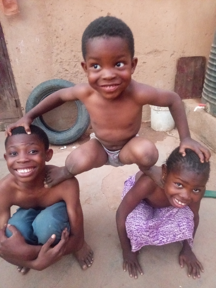 Three young children are playfully posing outdoors, likely in a yard or courtyard. The central figure is the youngest child, shirtless and wearing light grey underwear, squatting in the middle atop the other two. This child has a wide, joyful smile, looking upwards and slightly to the left, with both hands resting gently on the heads of the children below.
To the left, a slightly older boy is crouching or squatting, shirtless, with dark blue shorts or trousers bunched around his knees. He is holding his knees with his hands, smiling broadly at the camera. One foot of the child on top is placed on his shoulder or upper back.
To the right, a girl is in a kneeling or crawling position, wearing a purple patterned cloth or skirt. She is also smiling widely at the camera, and one of the top child's hands rests on her head. All three children have dark skin tones, short dark hair, and are barefoot, indicating a casual and comfortable setting. Their expressions convey happiness and a sense of fun.
The background consists of a light brown, earthy wall, possibly made of mud brick or stucco. Leaning against this wall behind the children is an old, dark tire. To the right, further back, there's a white bucket and a large, dark plastic barrel or container, possibly for water storage, with a small red spigot visible on its side. A dark wooden plank or board is also propped against the wall. The ground beneath the children is a light tan, suggesting dirt or concrete. The lighting appears natural and bright, indicating it is daytime. No text is visible in the image.