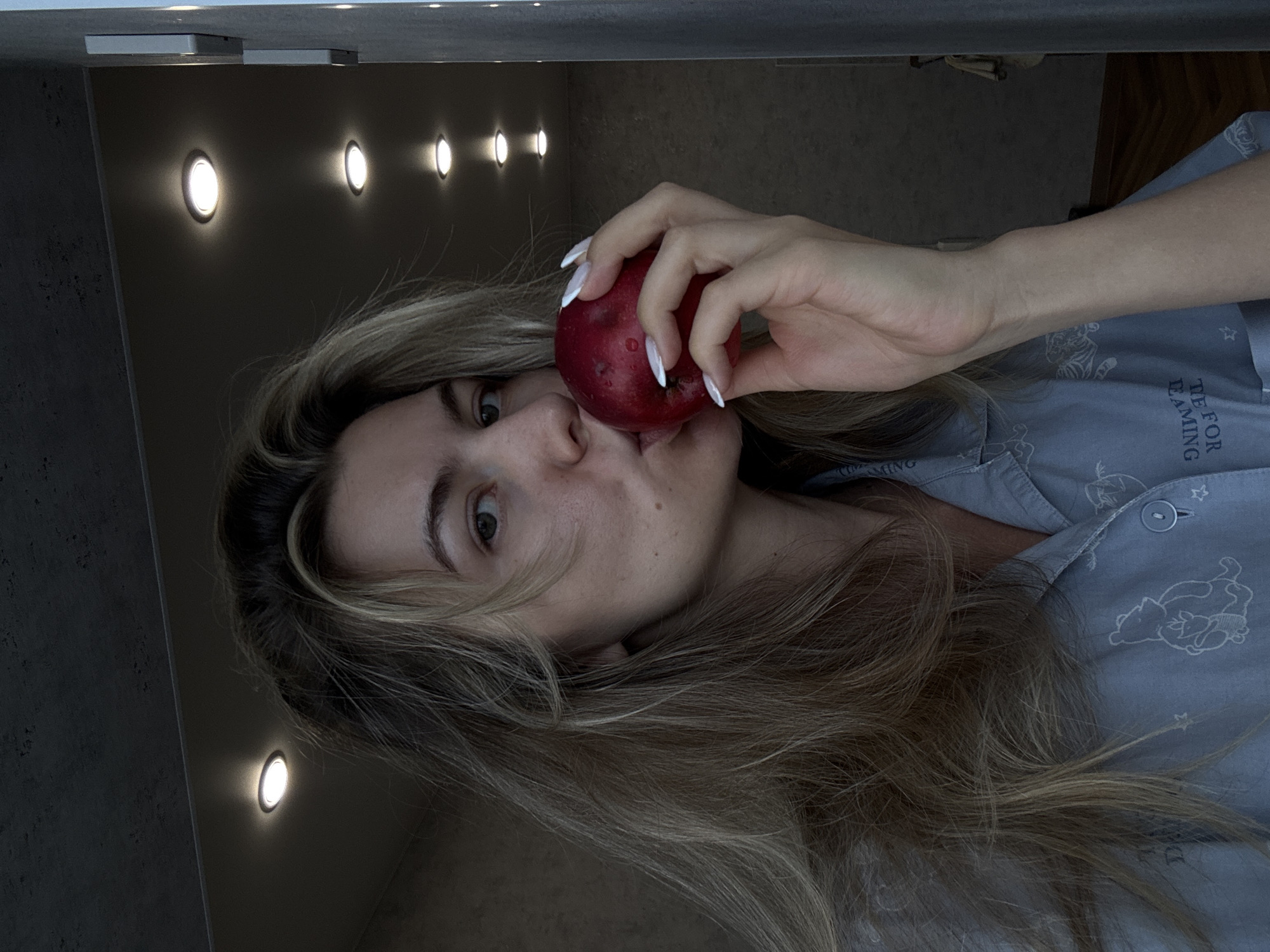 A young woman is pictured in an unusual, rotated perspective, appearing to be lying down and looking upwards towards the camera. She holds a red, unpeeled apple to her mouth, seemingly about to take a bite or having just done so. Her head is positioned to the left side of the frame, with her body extending to the right.
The woman has long, wavy, light brown or blonde hair, some strands of which frame her face. She has light-colored eyes and a neutral expression, looking directly at the viewer. Her left hand, adorned with long, white, almond-shaped manicured nails, holds the apple. She is wearing a light blue, collared pajama-style top with buttons down the front. The fabric features faint white cartoon illustrations, including what appears to be Winnie the Pooh and Piglet, along with small stars. Partially visible text on her top includes "TE FOR" and "REAMING," suggesting the phrase "TIME FOR DREAMING."
The background consists of dark, textured surfaces, likely a wall and a ceiling or an overhead shelf. Several circular, bright white recessed lights illuminate the dark background, creating a line of glowing orbs stretching into the distance. Two rectangular, lighter-colored objects, possibly light fixtures or other fixtures, are also visible attached to the overhead surface near the top left corner of the frame. The overall setting appears to be indoors, possibly a bedroom or a cozy living space, with artificial lighting providing the illumination.