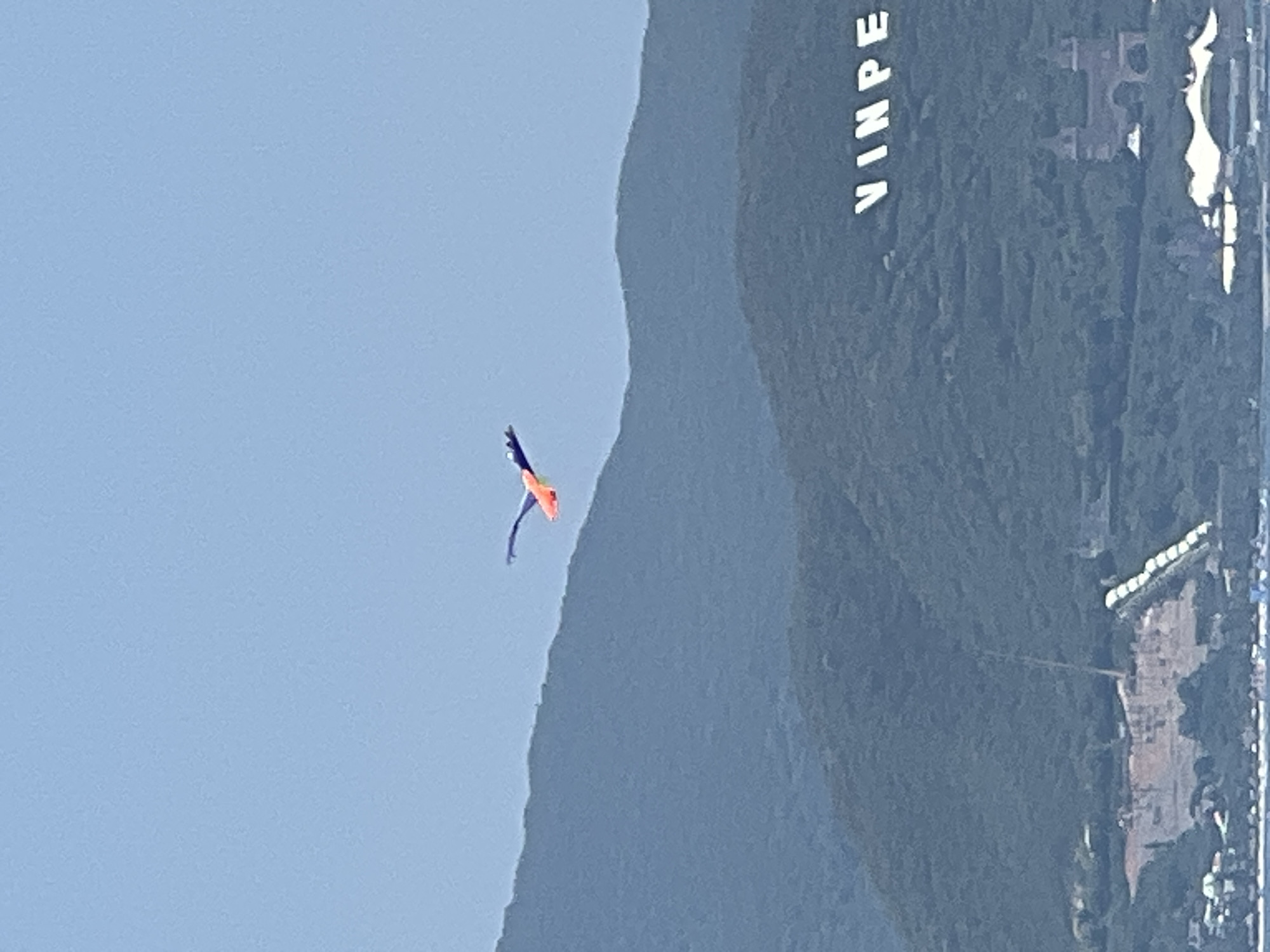 The image captures a scenic view of Nha Trang, Vietnam, with a kite shaped like a bird soaring through a clear blue sky. The kite, predominantly orange and blue, is in mid-flight, its wings outstretched. Below, verdant, tree-covered hills form a dramatic backdrop. In the foreground, the large white letters "VINPE" are visible, hinting at a location or landmark. Further down the hillside, a large structure resembling a castle can be seen, along with what appears to be a resort or hotel complex. The lower portion of the image shows a body of water, likely the sea, with some structures or boats. The scene suggests a bright, sunny day.