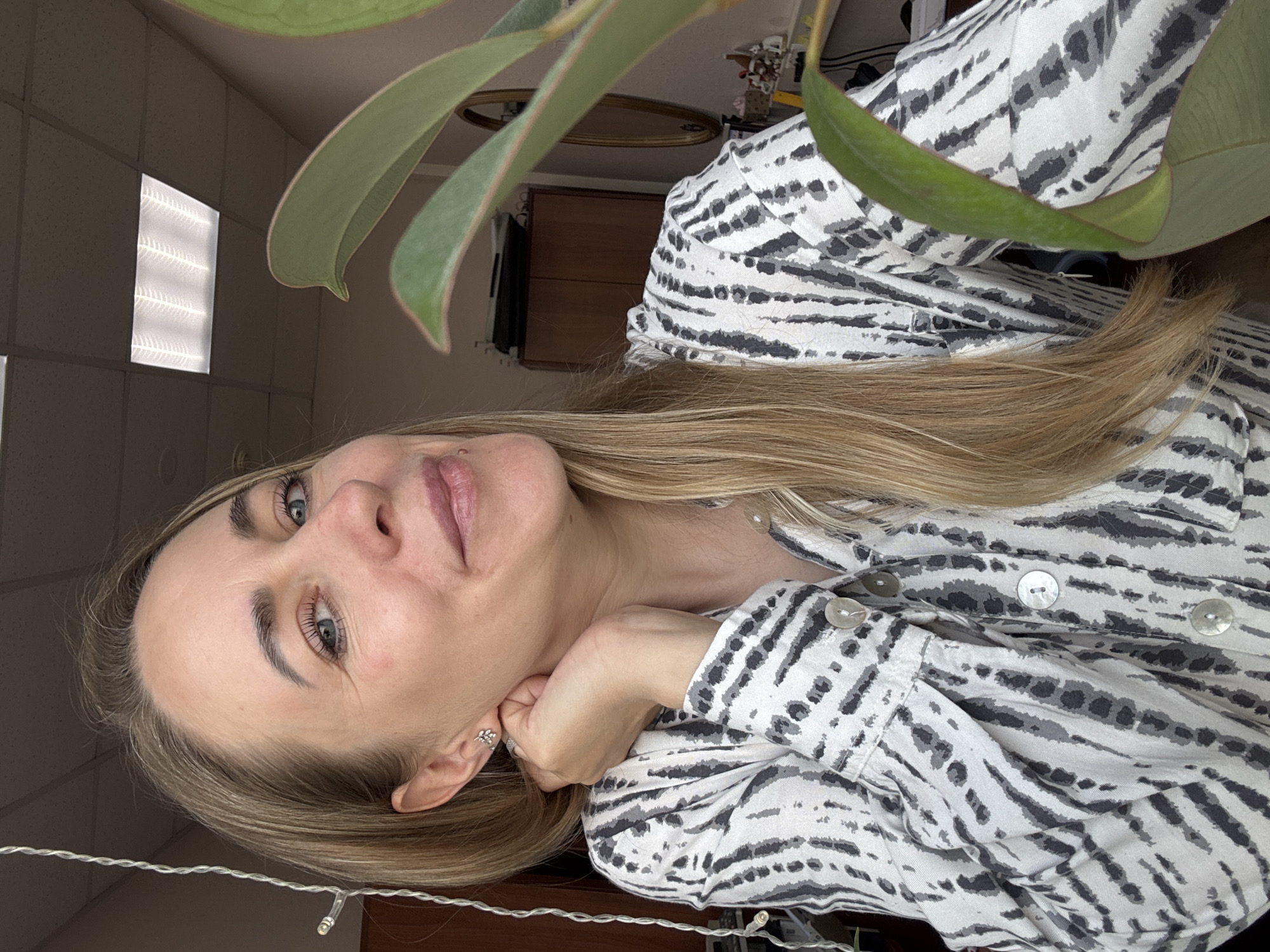 A Caucasian woman with blond hair and blue eyes is centered in the frame, resting her chin on her hand. She wears a light-colored button-down shirt with a grey-and-white zebra stripe pattern. A green houseplant with large leaves partially obscures the upper portion of the frame. The background shows an interior space with a row of fluorescent ceiling lights and dark brown furniture. A string of lights hangs in the lower portion of the frame. Based on the environment details and the absence of specific landmarks, it is not possible to conclusively confirm the location as Chaikovsky, Russia. Further visual or contextual cues would be required to make that determination.