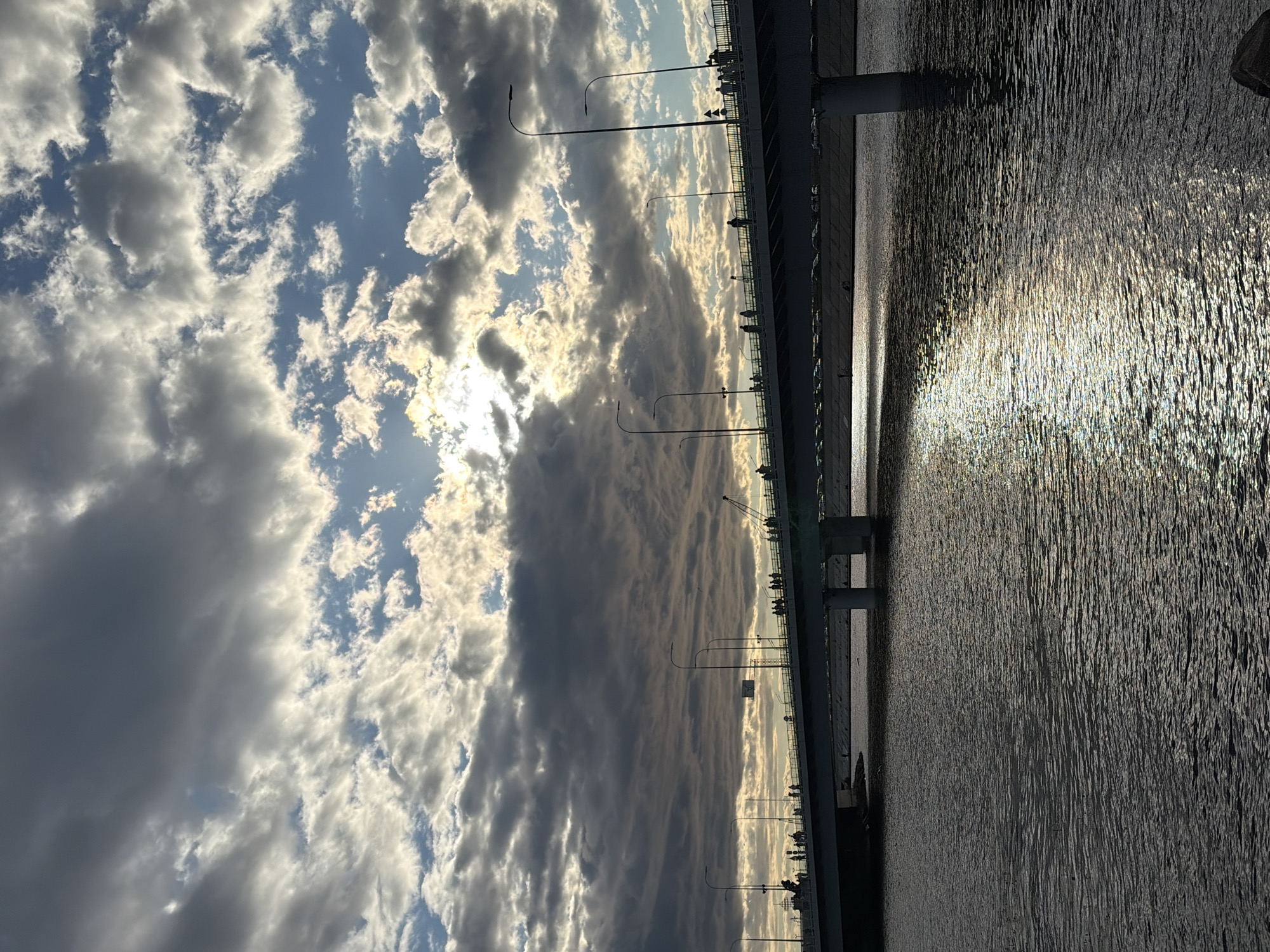 A wide-angle shot captures a bridge crossing a body of water in Saint Petersburg, Russia. The bridge, a prominent feature, is lined with streetlights and populated by numerous silhouetted figures, suggesting pedestrians enjoying the view or commuting. The water below reflects the dramatic sky, which is filled with a mix of dark, heavy clouds and brighter patches where sunlight breaks through, creating a shimmering effect on the surface. The overall scene evokes a sense of a cool, overcast day with intermittent sunshine.